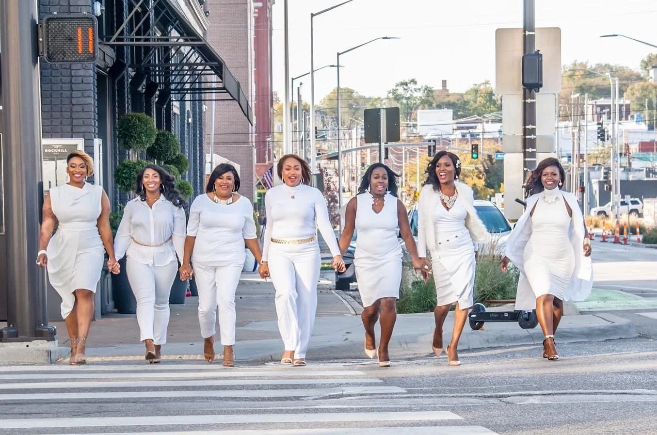 Group of hairstylists dressed in white walking across a city crosswalk, smiling and holding hands, with cars and streetlights in the background for a branding session