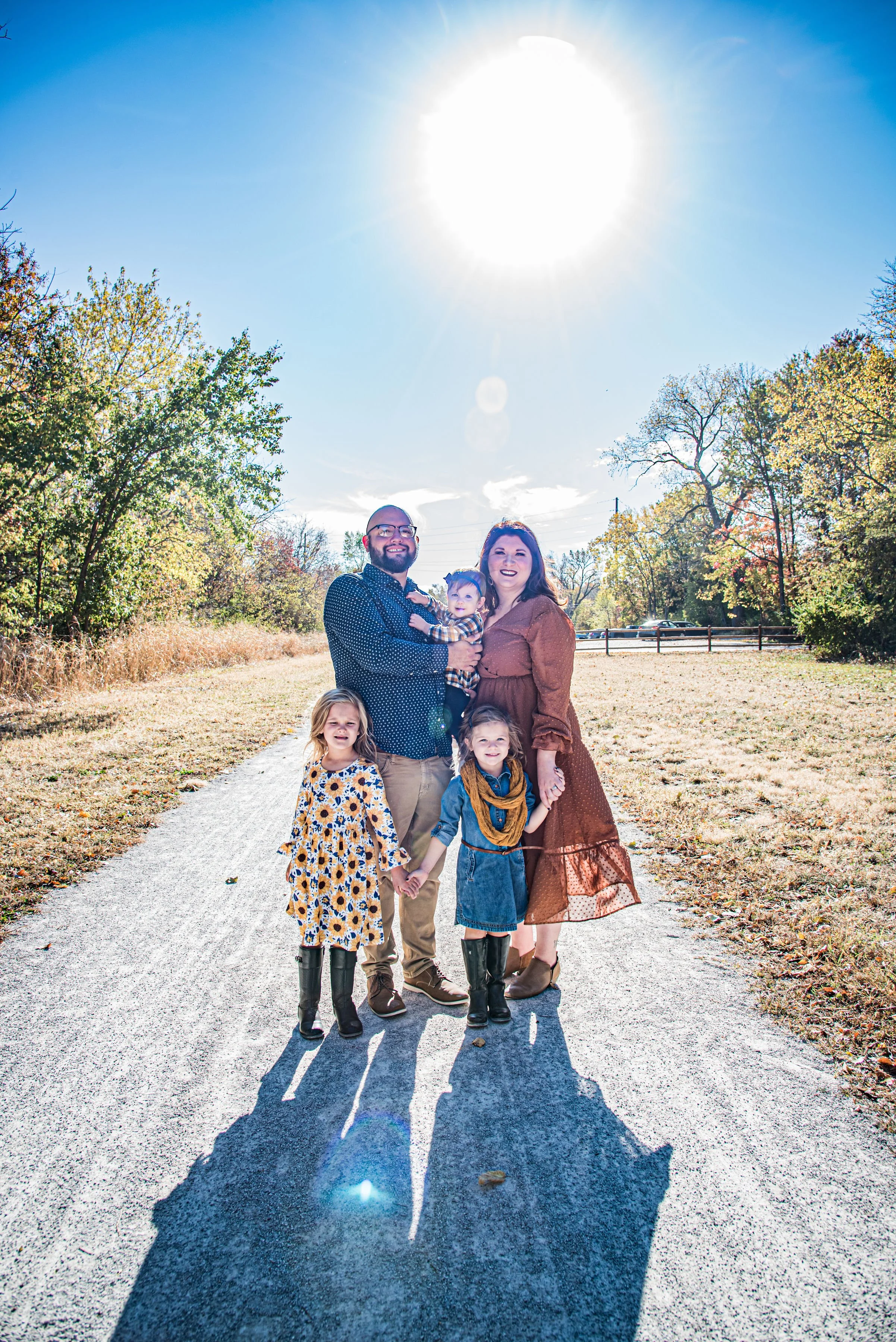 A family of six standing on a gravel pathway outdoors under the bright sun, with trees and a fence in the background. The group includes a man, a woman, two girls, a boy, and a toddler, all smiling and holding hands.