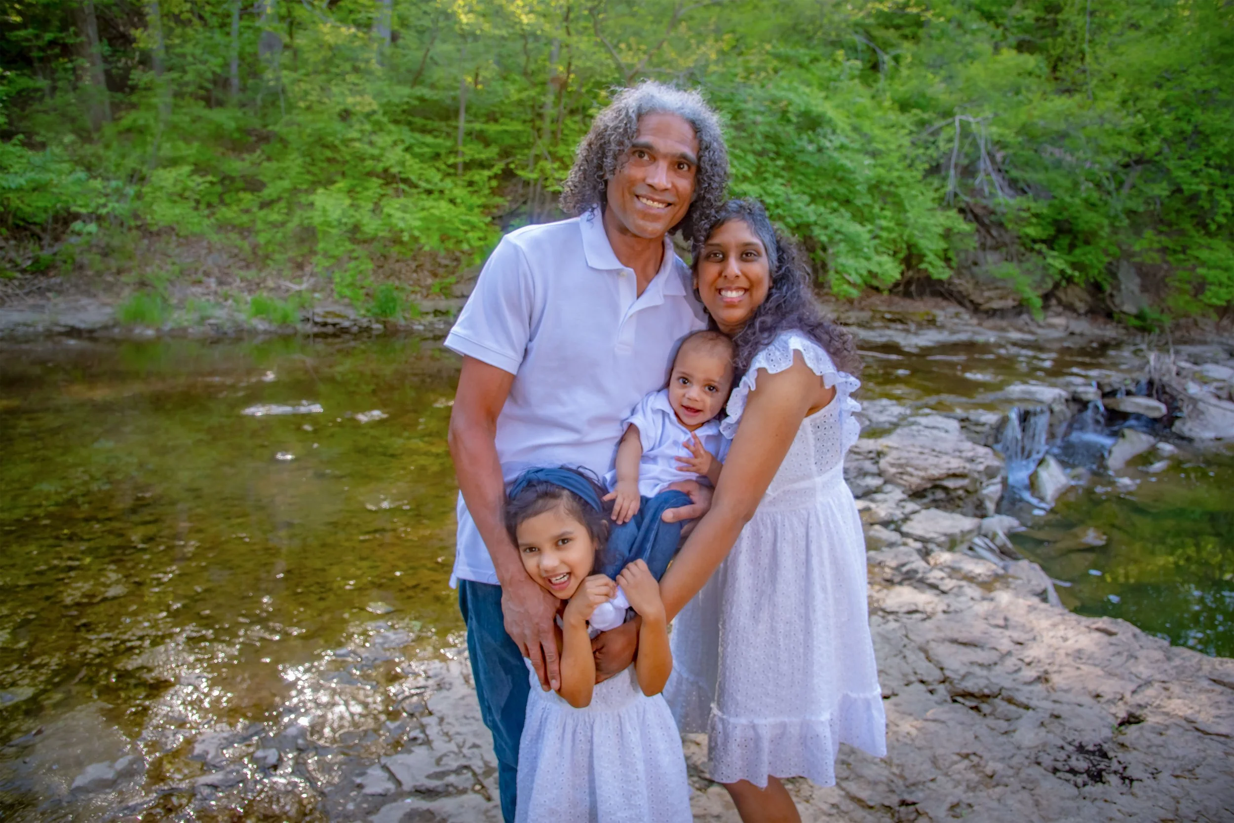 A family of four standing together by a river in a green wooded area. The father, mother, and two young girls are smiling at the camera. The mother is holding one of the girls, and the other girl stands in front of the father.