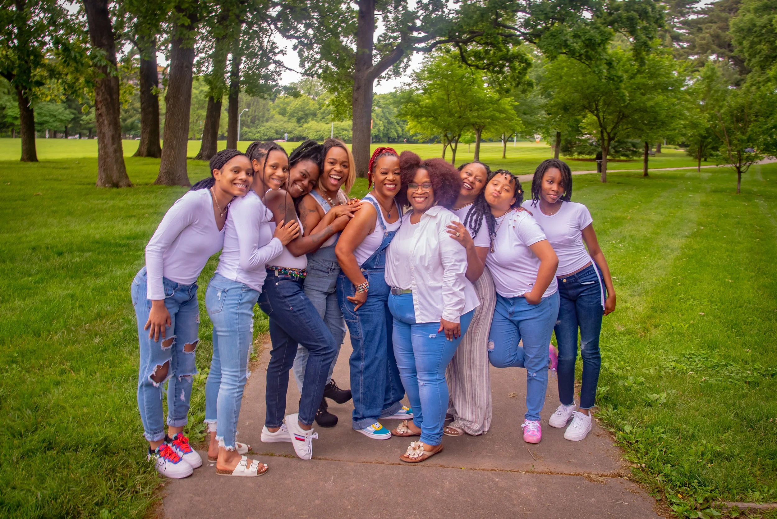 Group of women and girls standing on a sidewalk in a park with trees in the background, smiling and embracing.