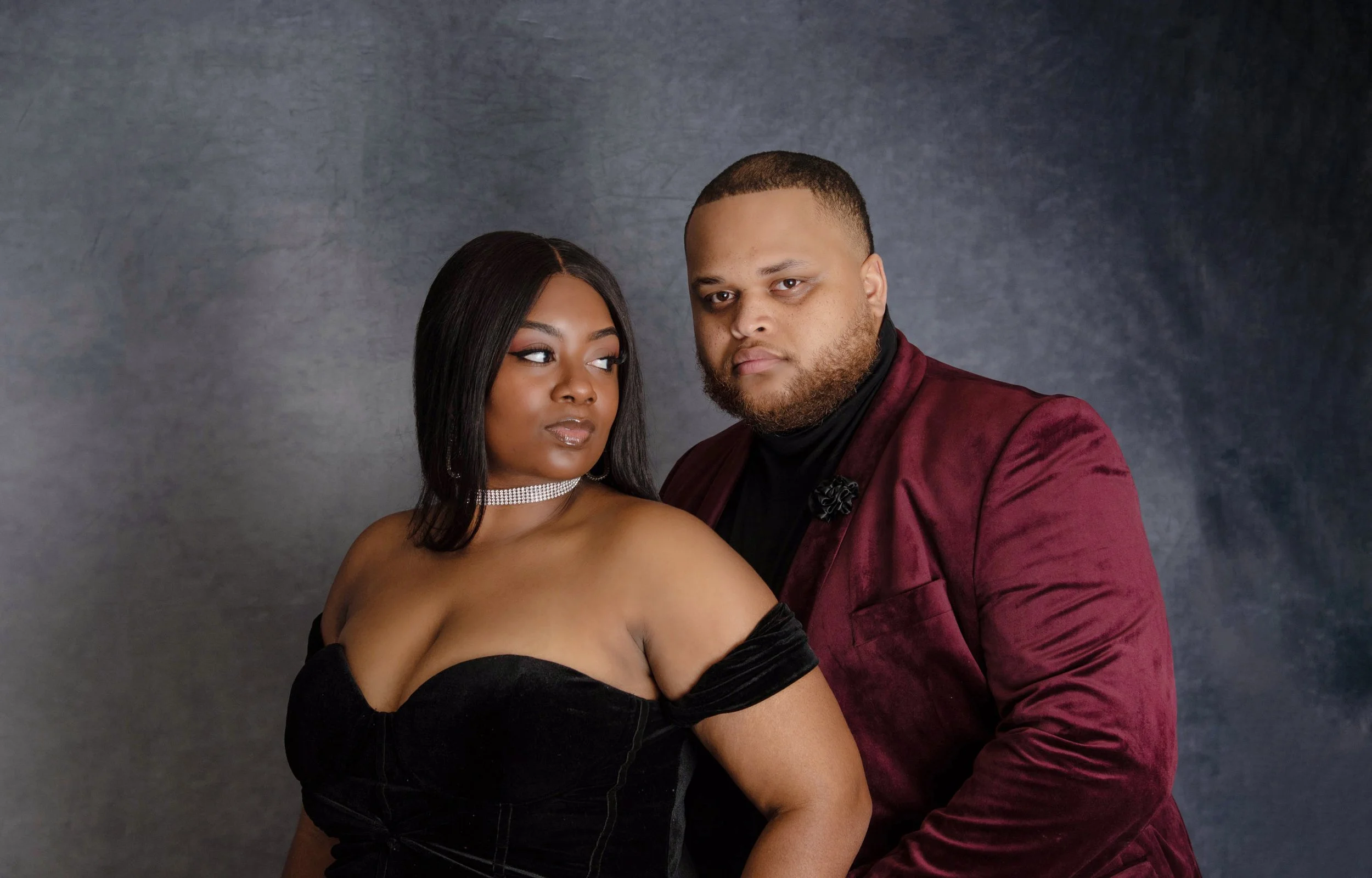 A couple posing for a portrait photograph against a dark gray background. The woman is wearing a black strapless dress with off-the-shoulder sleeves and a rhinestone choker, and has shoulder-length black hair. The man is dressed in a burgundy blazer 