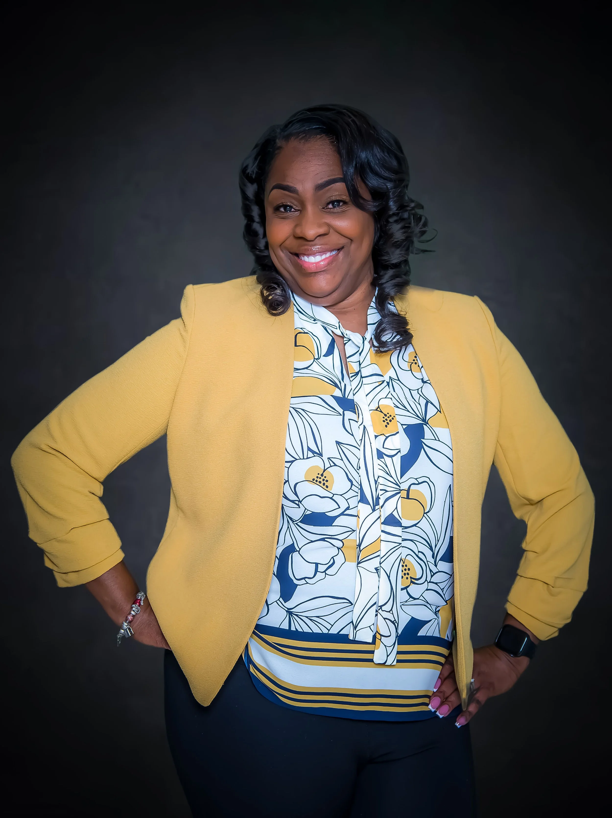 A woman smiling with hands on her hips, wearing a yellow blazer over a floral blouse against a dark background for branding session.