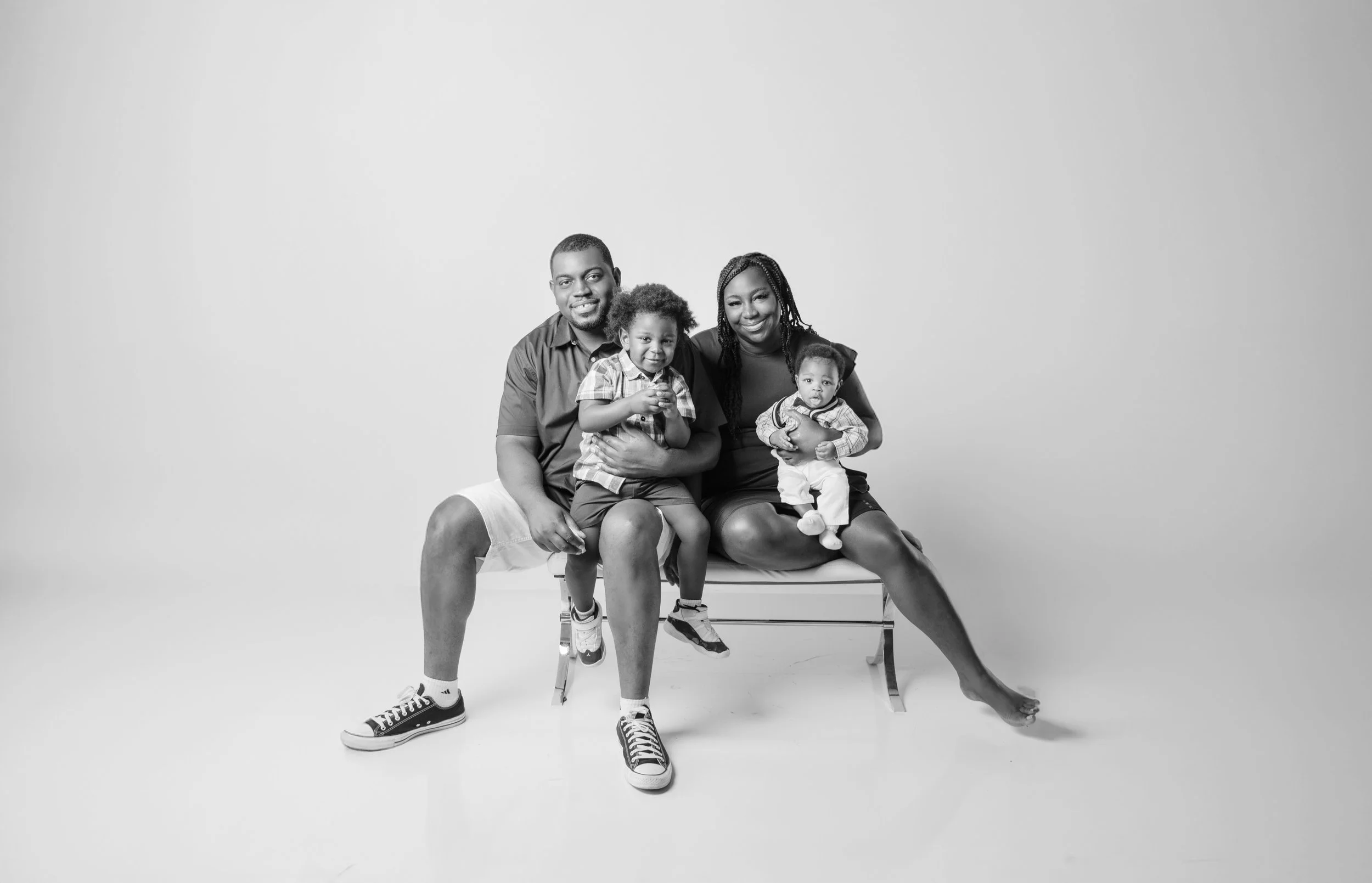 Black and white photo of a family of four sitting on a bench against a plain background. The father is on the left, smiling, with his arm around a young boy sitting on his lap. The mother is on the right, also smiling, holding a baby girl.