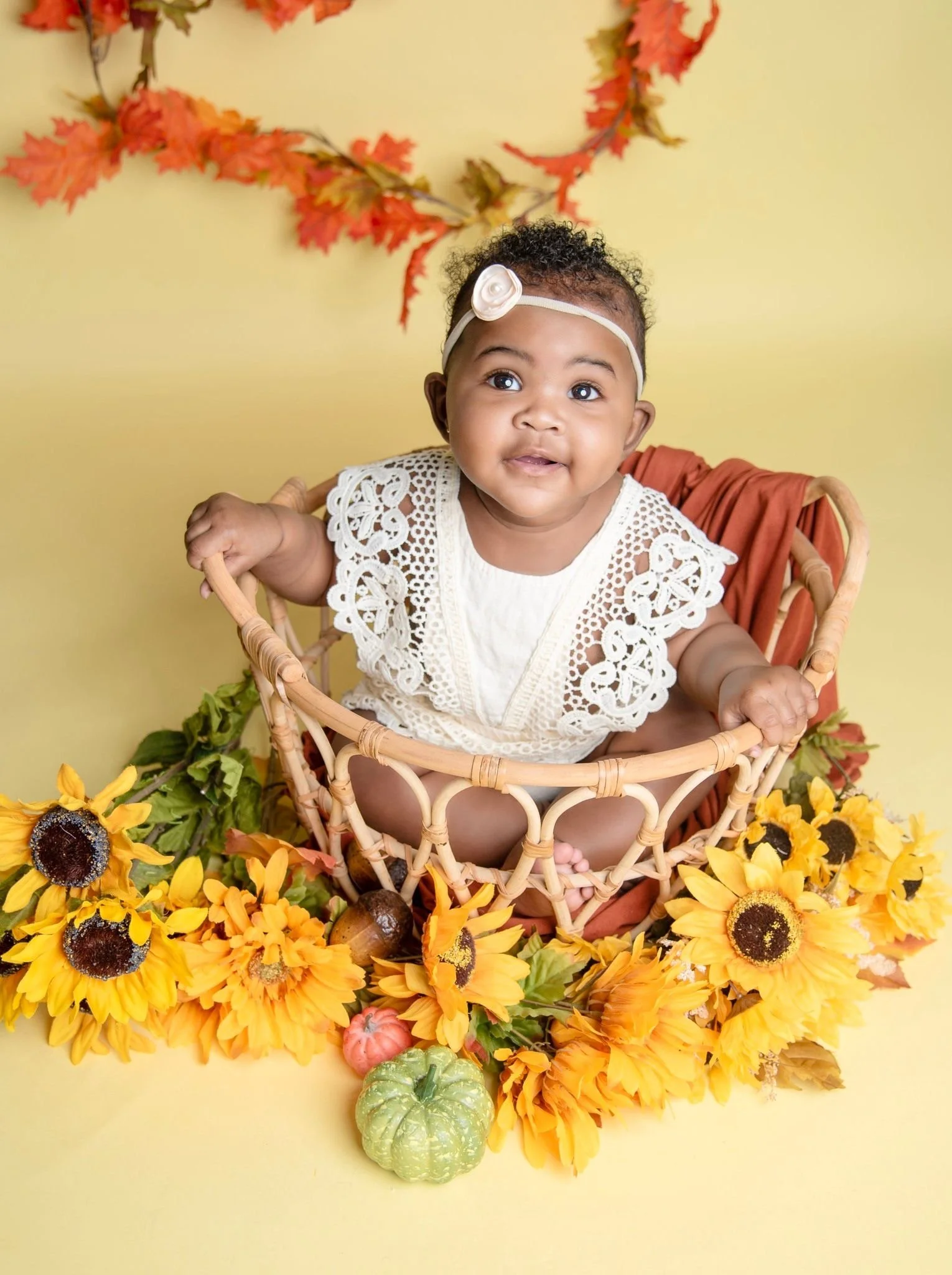 Adorable baby girl with curly hair wearing a white lace dress and a white headband with a flower sitting in a wicker basket surrounded by yellow sunflowers, small pumpkins, and autumn leaves with a yellow background.