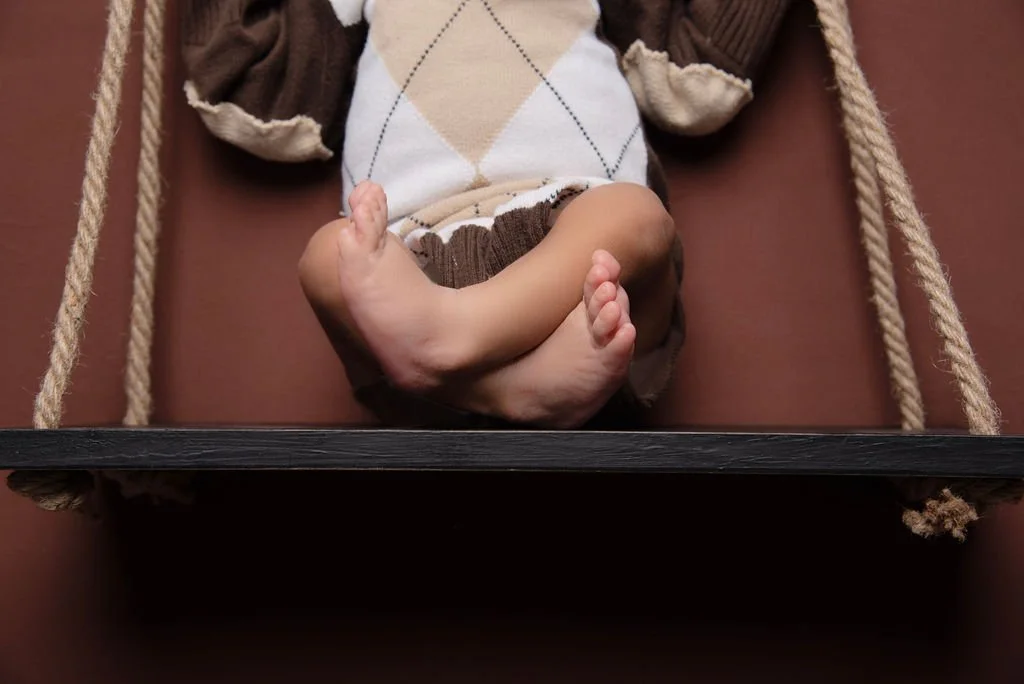 Baby lying in a wooden swing with a brown cushion and beige ropes, wearing a sweater and shorts.