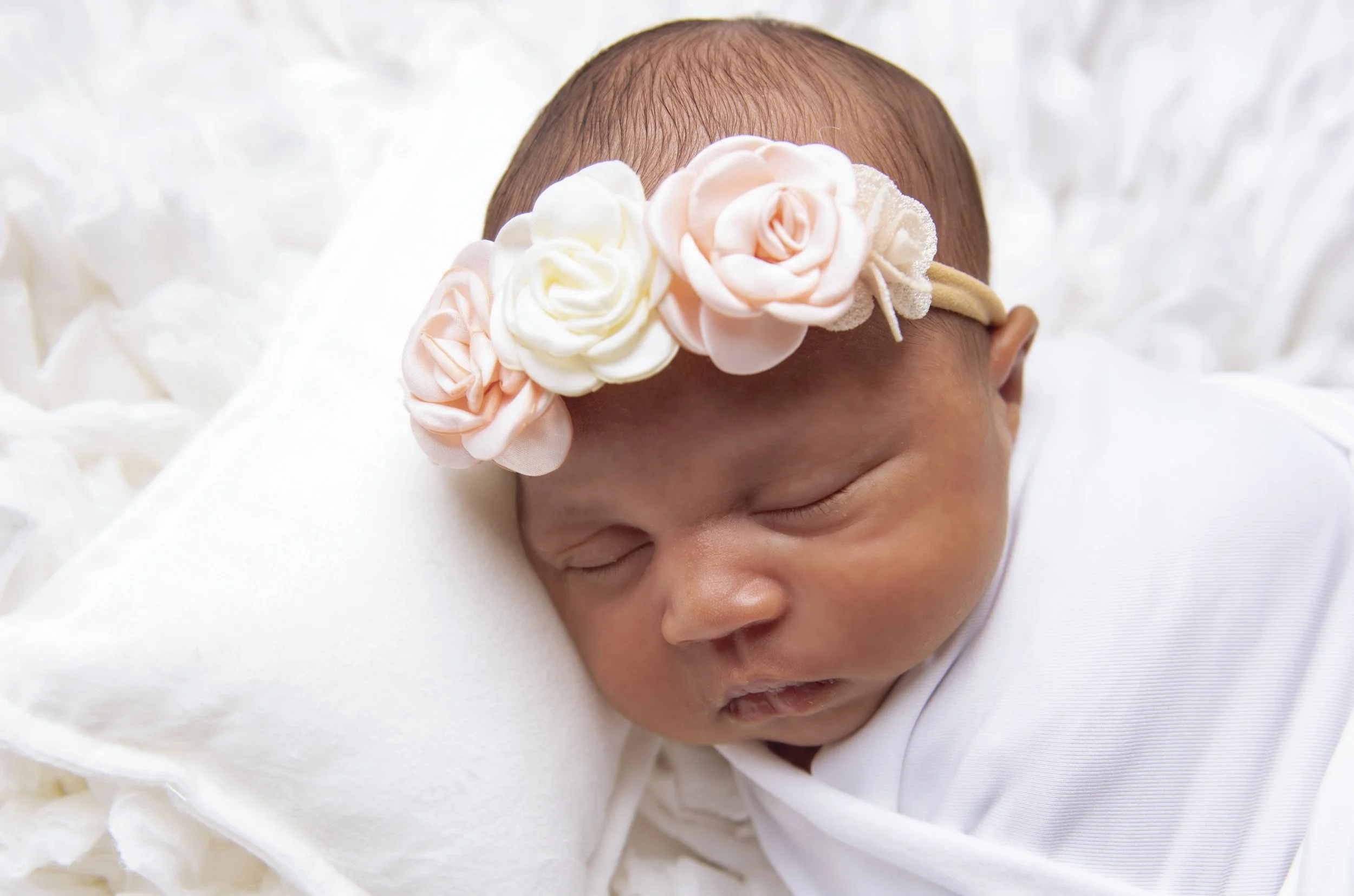 Close-up of a sleeping newborn baby with a pink and white flower headband, resting on a soft white surface.