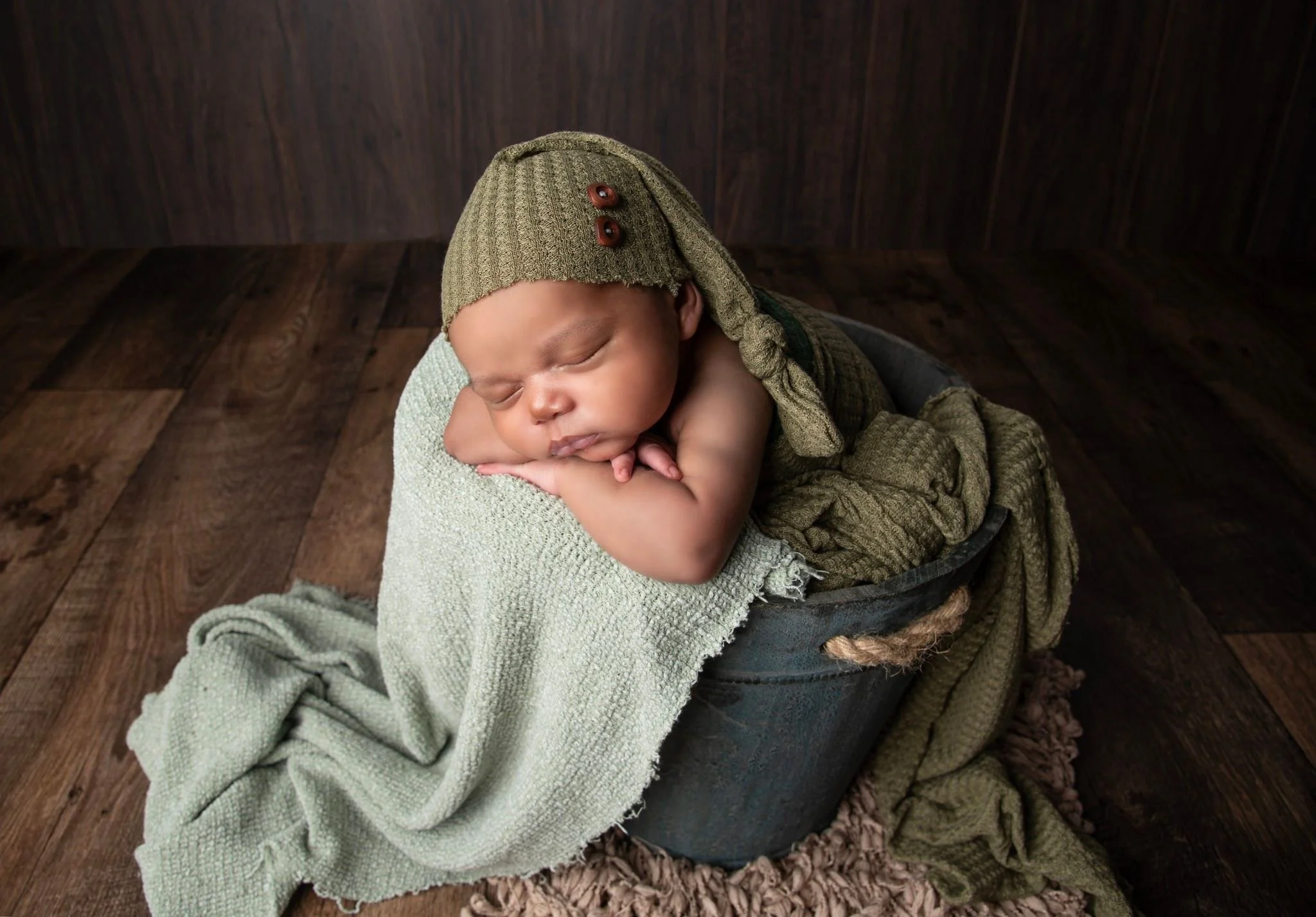 A newborn baby sleeping on folded blankets inside a black bucket, wearing a green knit hat with buttons, surrounded by cozy green and brown fabrics.