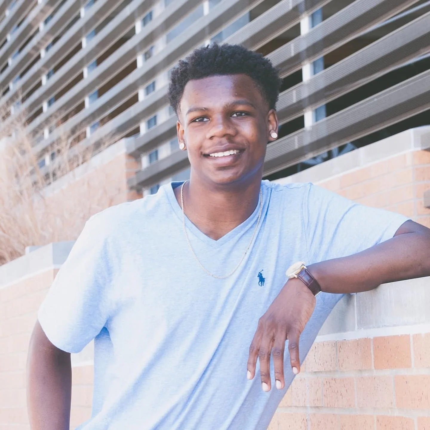 A young man with dark skin, curly black hair, and earrings smiling outdoors. He is wearing a light blue t-shirt with a Polo Ralph Lauren logo, a watch, a gold chain, and is leaning on a brick wall with one arm, in front of a modern building with hori
