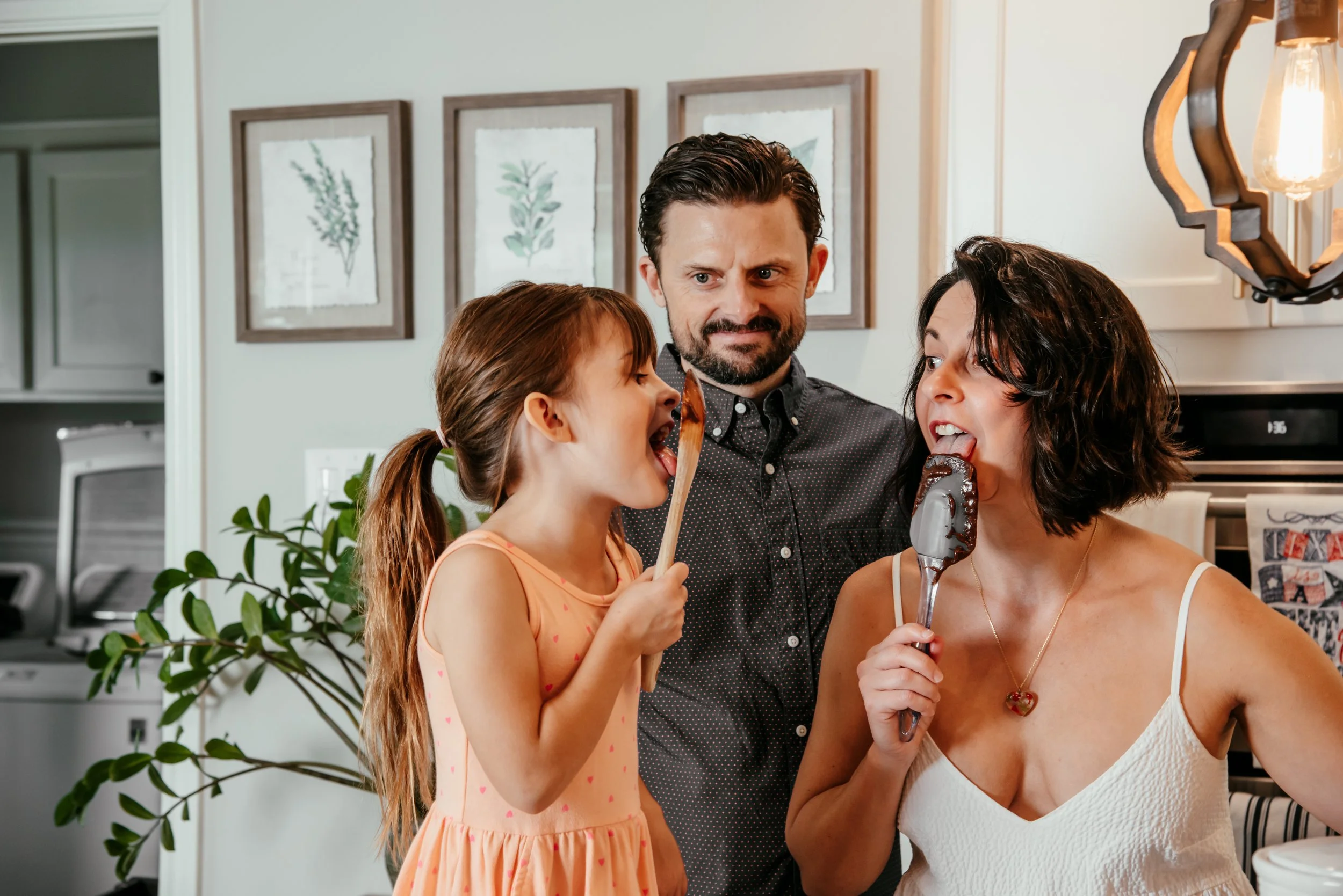 A family having fun in the kitchen, with a girl and woman licking ice cream while a man looks on.