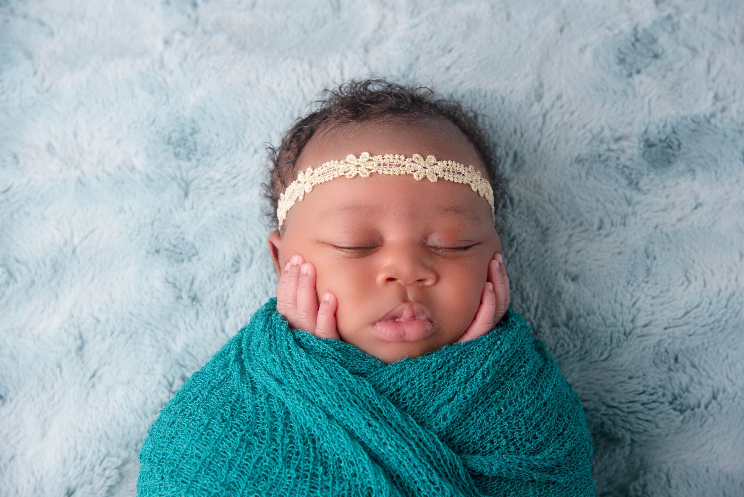 A sleeping baby with a lace headband and a teal wrap, resting on a fluffy light blue blanket.