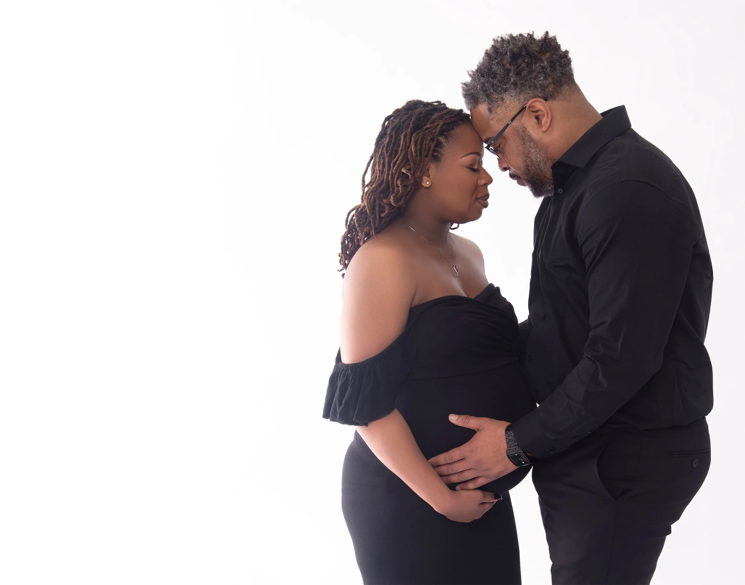 A pregnant woman and a man face each other with foreheads touching, both with eyes closed, in a tender moment against a plain white background.