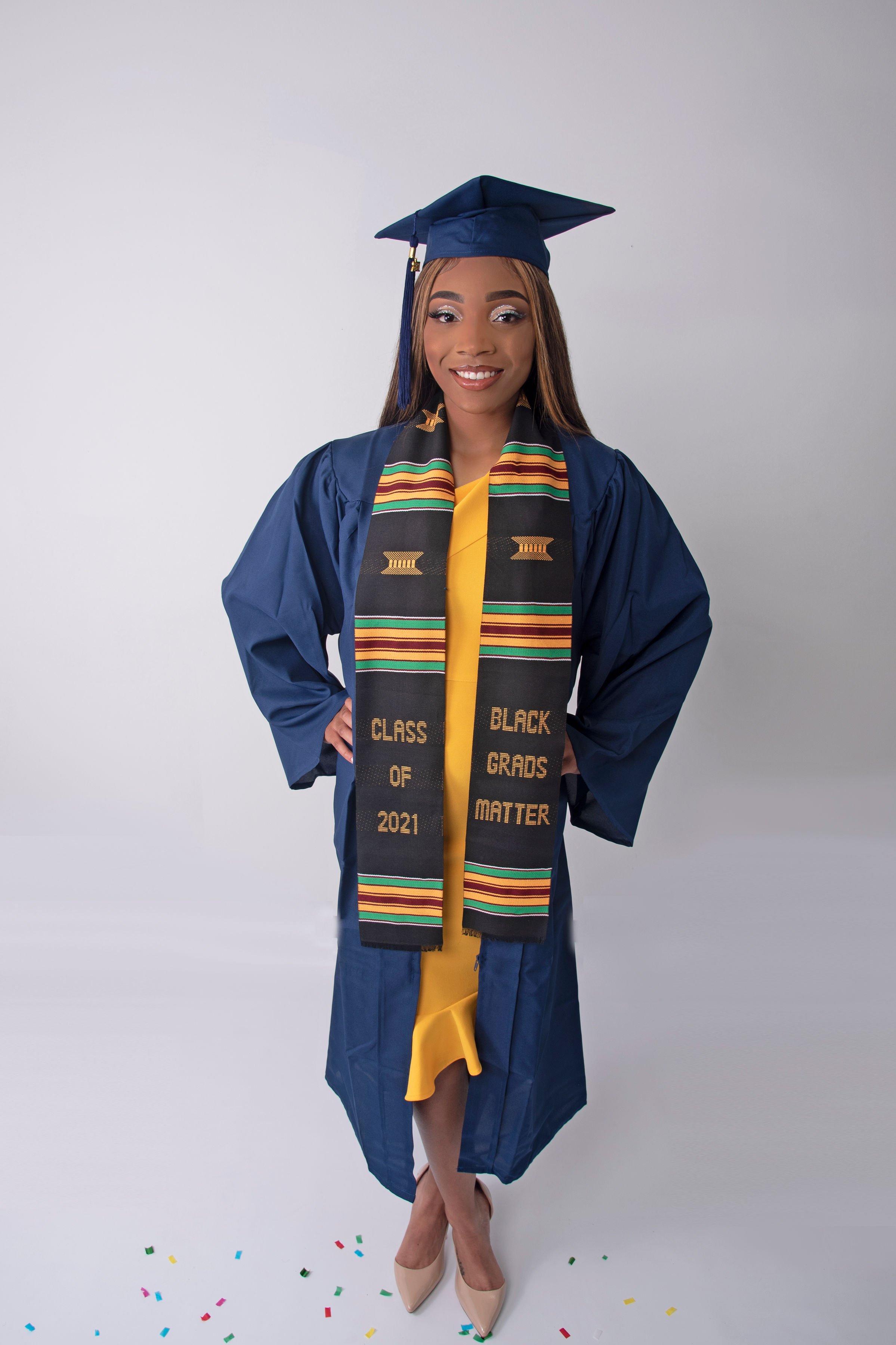 Young woman in blue graduation gown and cap, wearing a black kente cloth stole that says "Class of 2021, Black Graduates Matter," standing with hands on hips, smiling, with confetti on the floor.