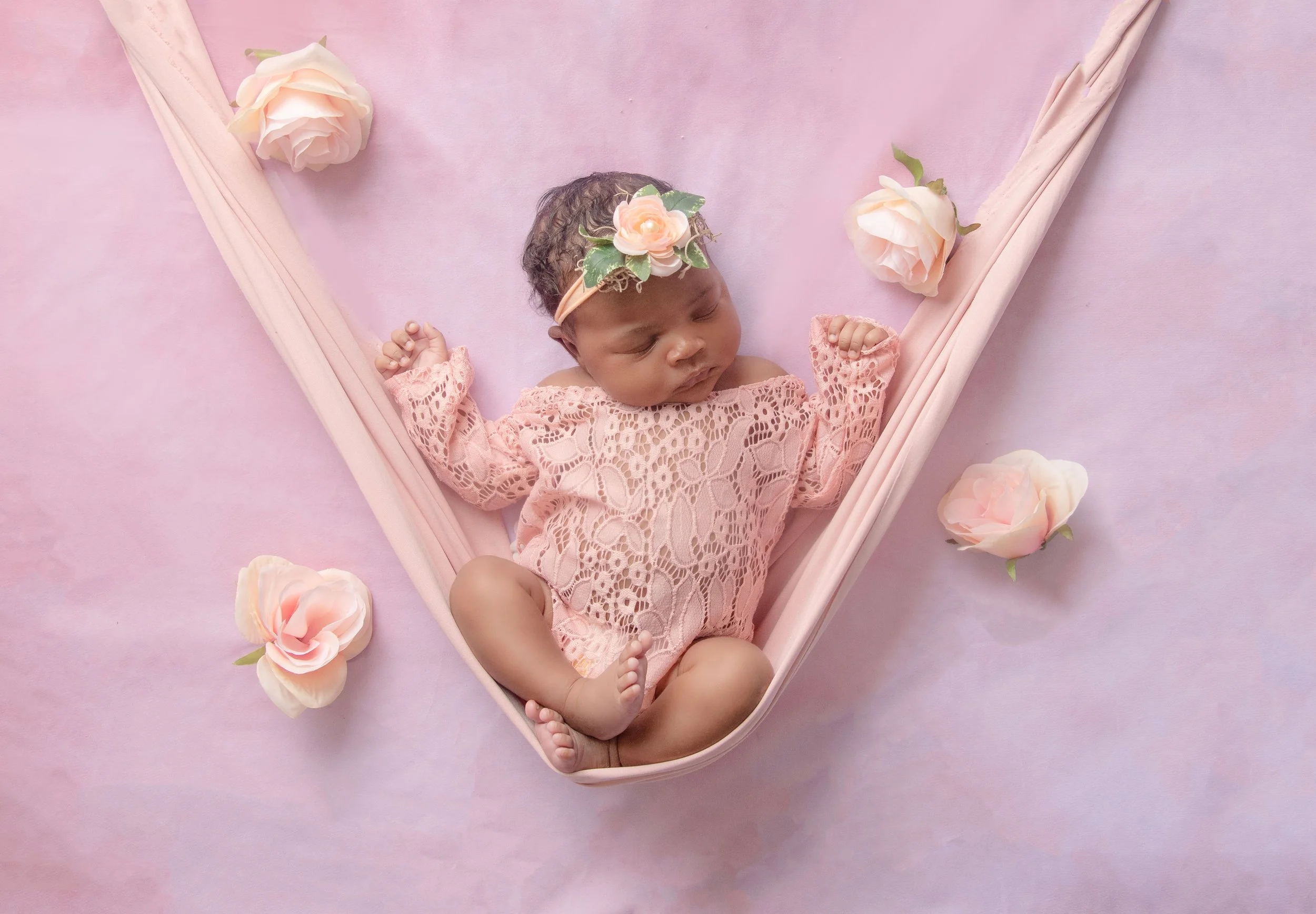 A sleeping baby girl wearing a pink lace dress and a floral headband, lying on a pink blanket surrounded by pink and white roses, with a pink fabric border forming a V-shape around her.