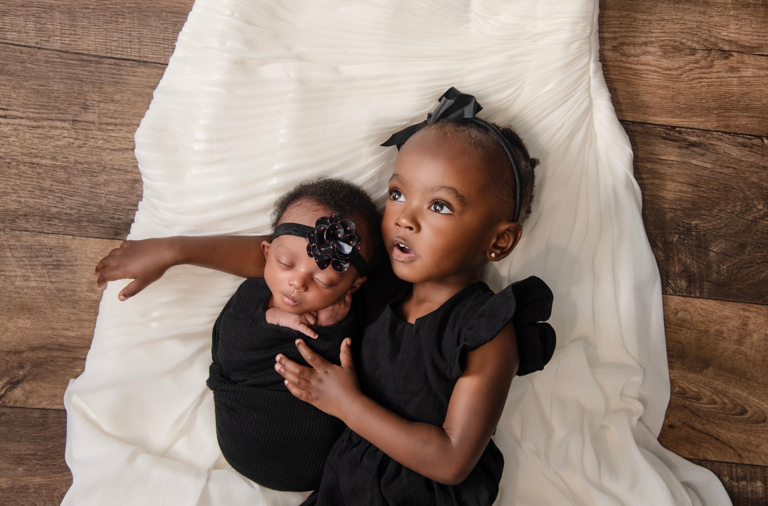 Two young girls with matching black headbands and black dresses lying on a white fabric on a wooden floor, one girl is awake and the other girl is sleeping.