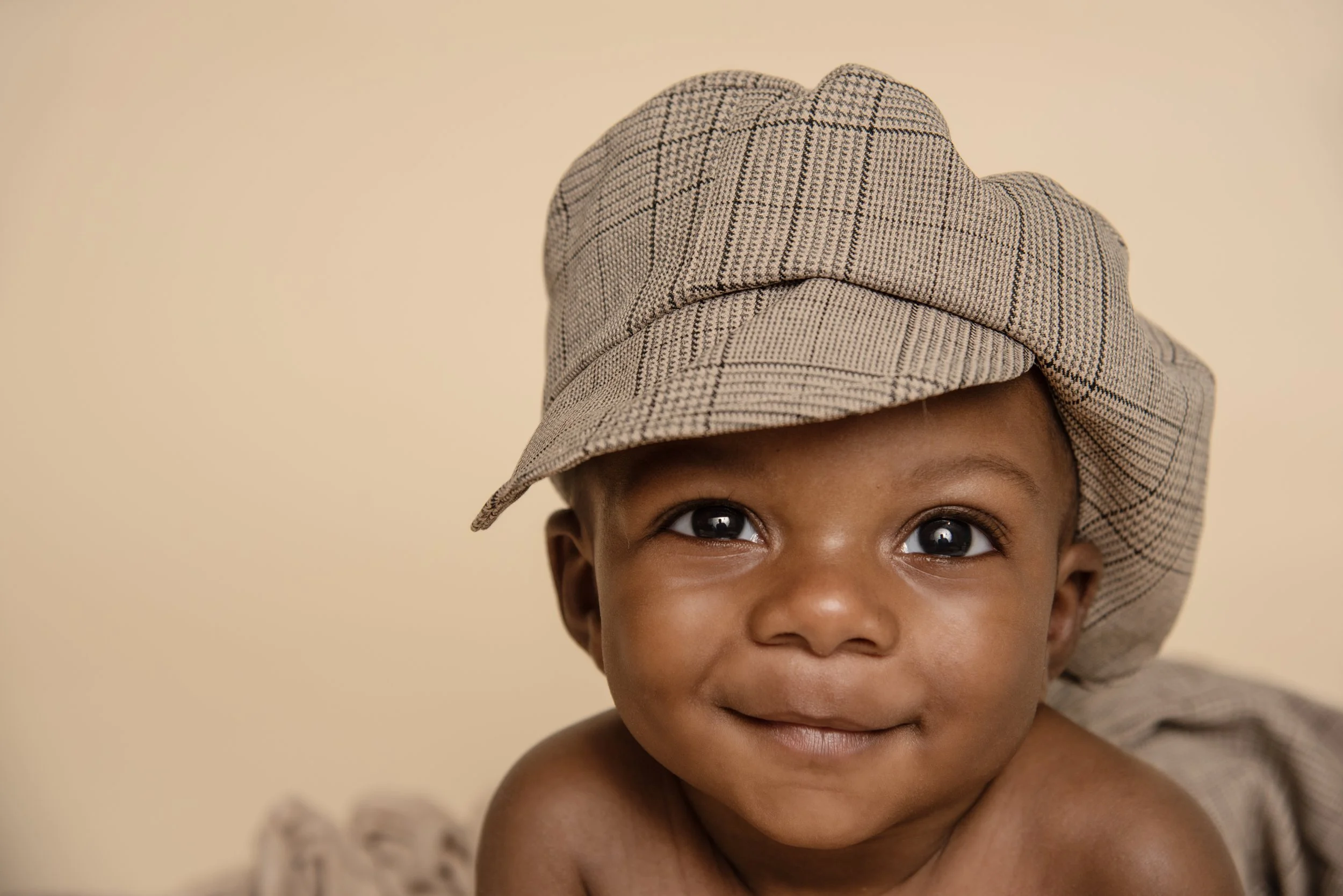 A young child with a brown complexion, smiling, wearing a tan plaid hat, lying on their stomach against a beige background.