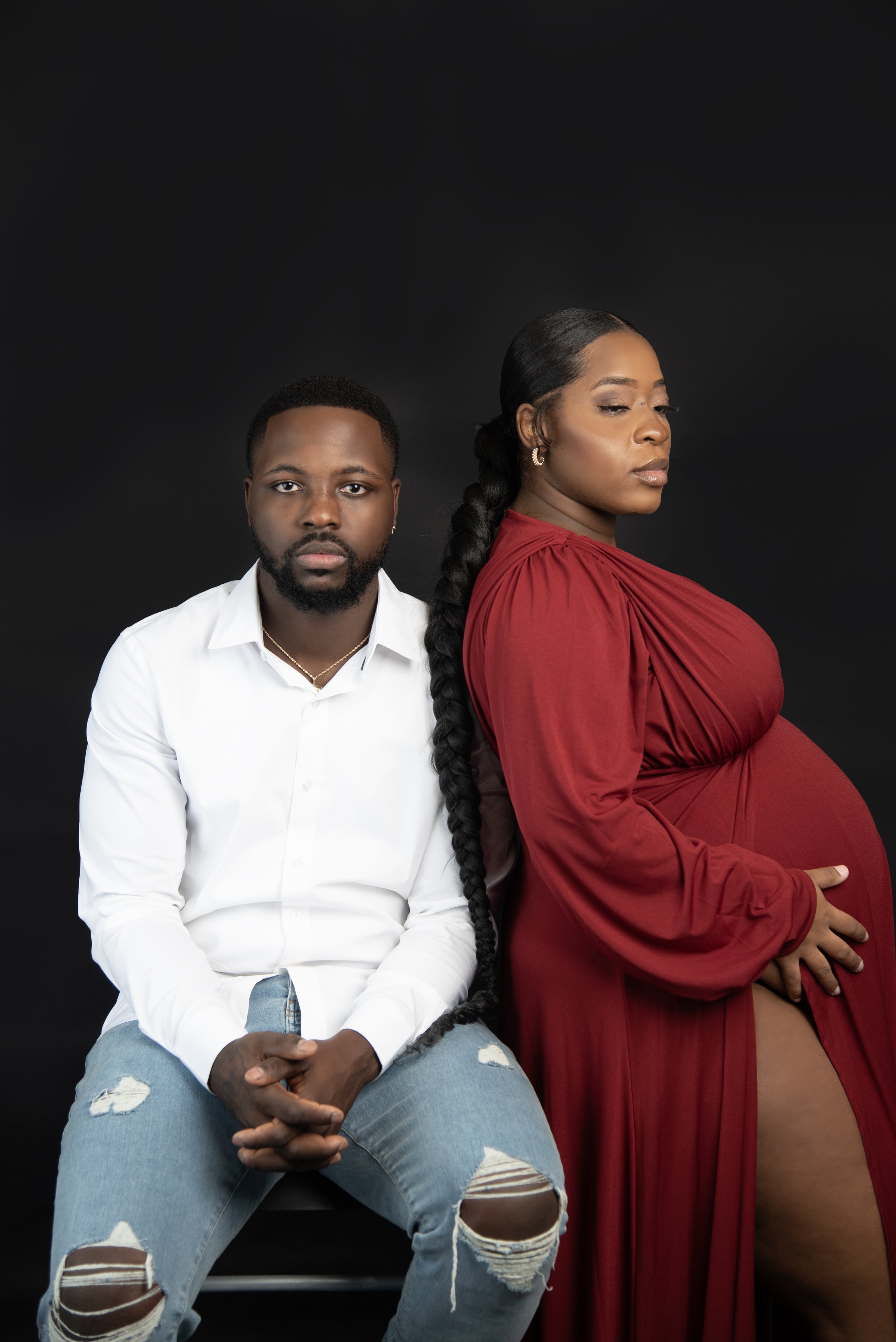 A man with a white shirt and ripped jeans sits with hands clasped, and a pregnant woman in a red dress stands close, holding her belly, against a black background.