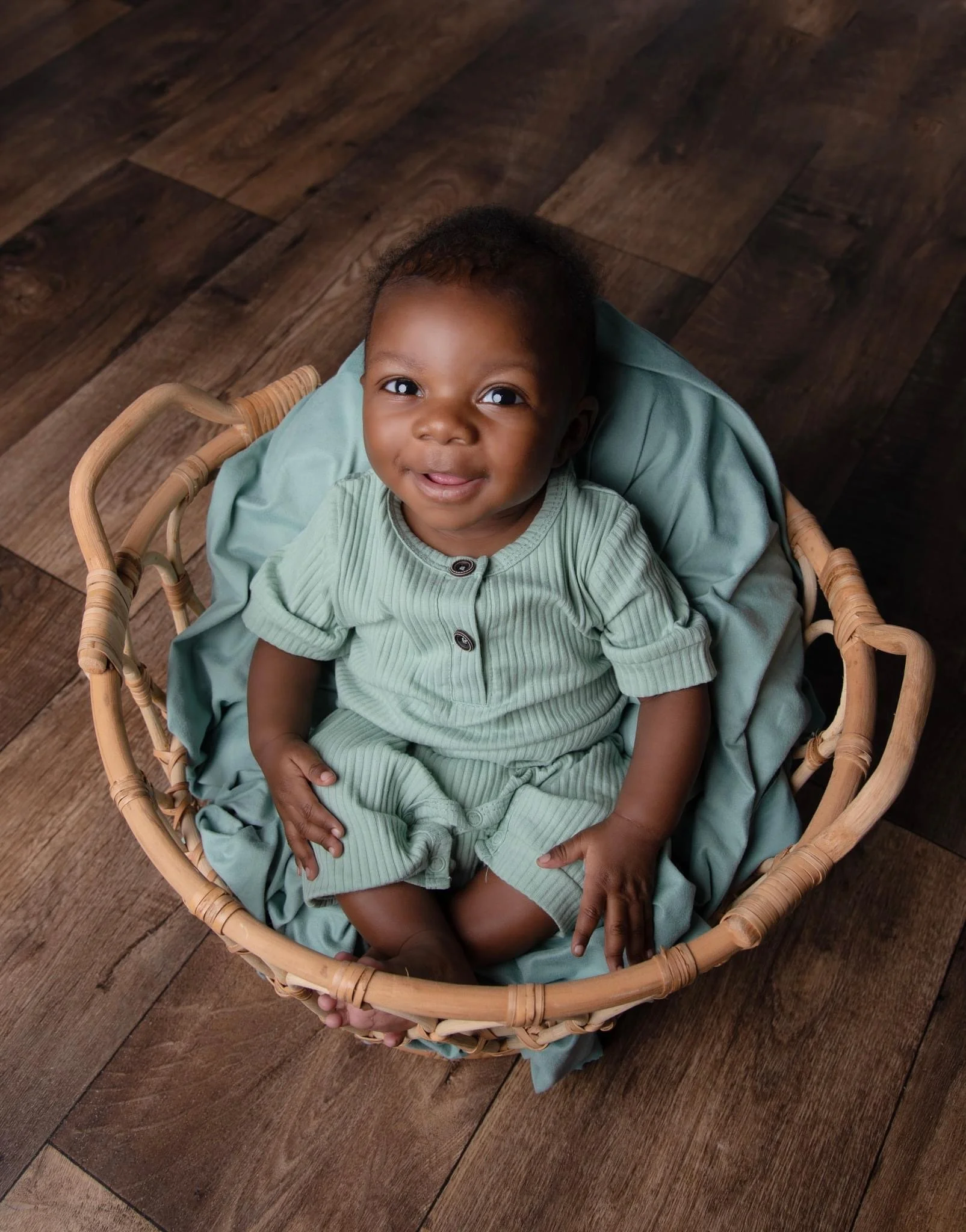 A smiling baby with dark skin and curly hair sitting in a wicker basket, wearing a green outfit, on a wooden floor.