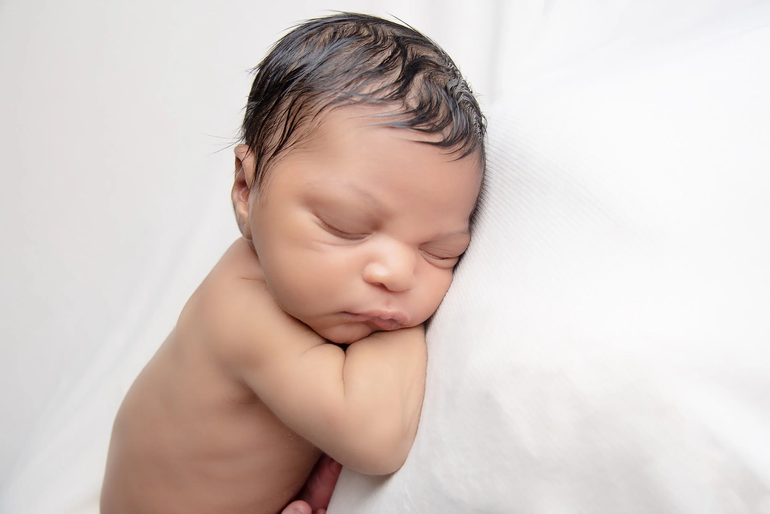 A sleeping newborn baby rests on a white surface with arms crossed under its face.