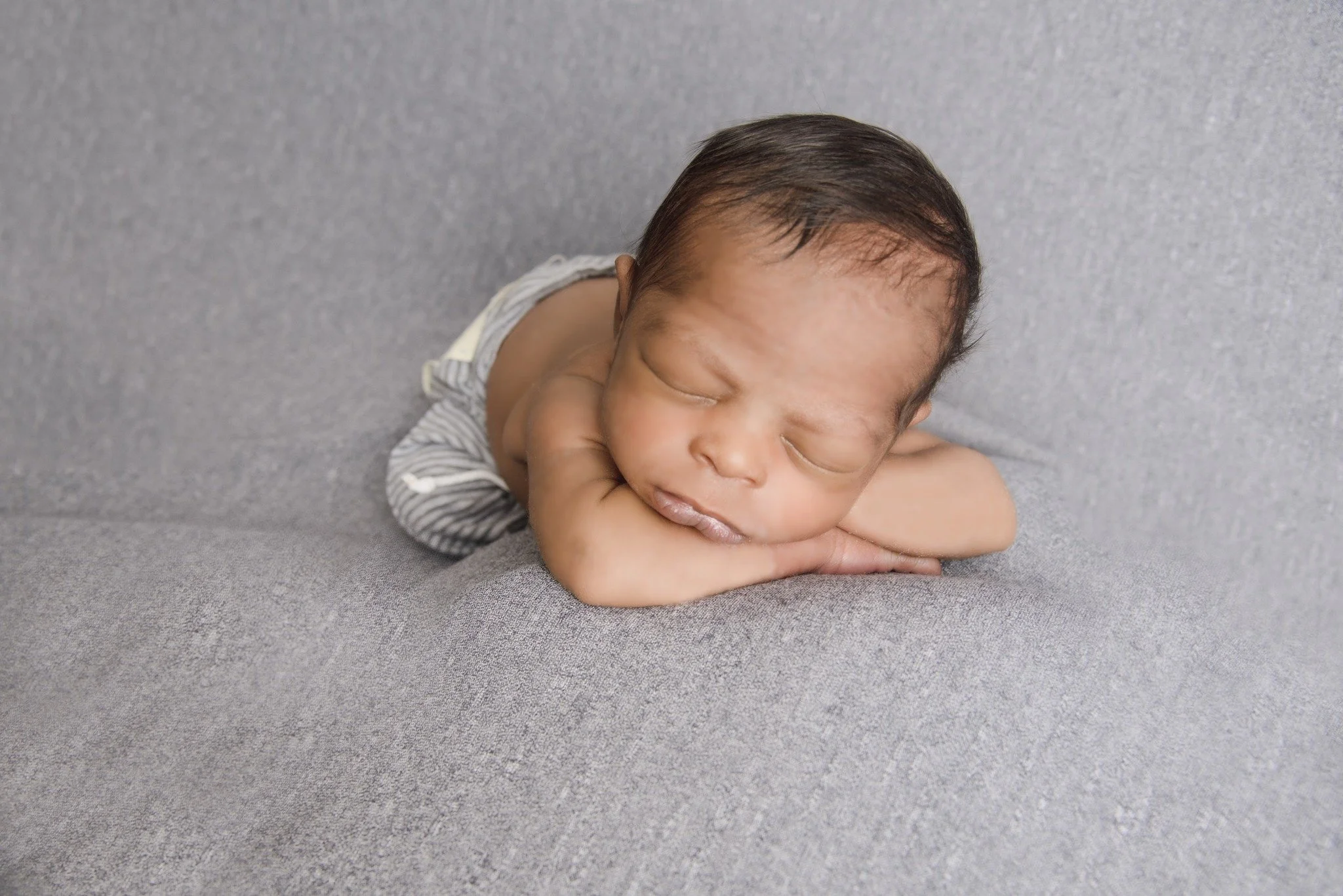 A newborn baby sleeping on a gray surface, with head resting on arms, wearing striped clothing.