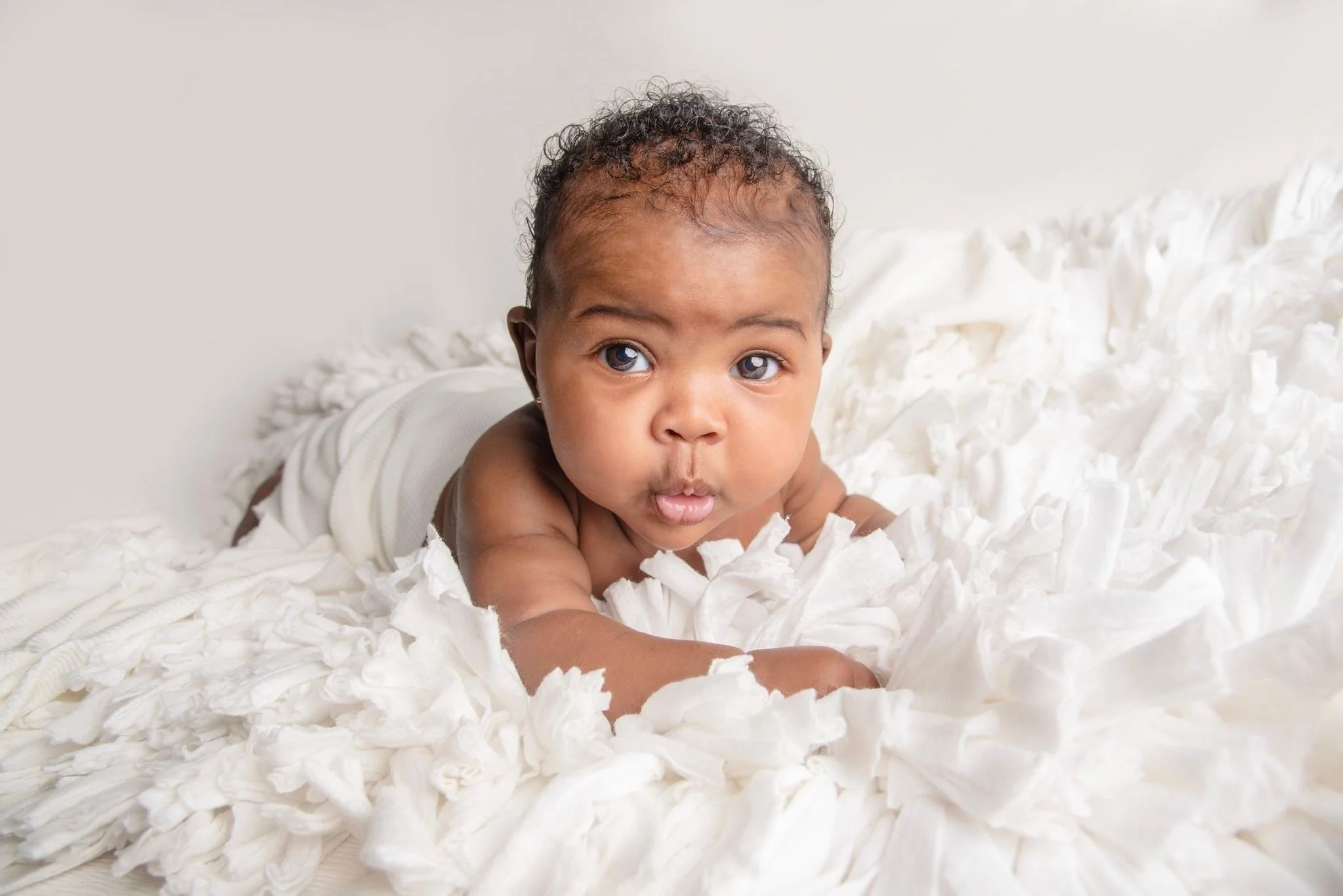 Baby lying on a white fluffy blanket, looking at the camera with wide eyes.