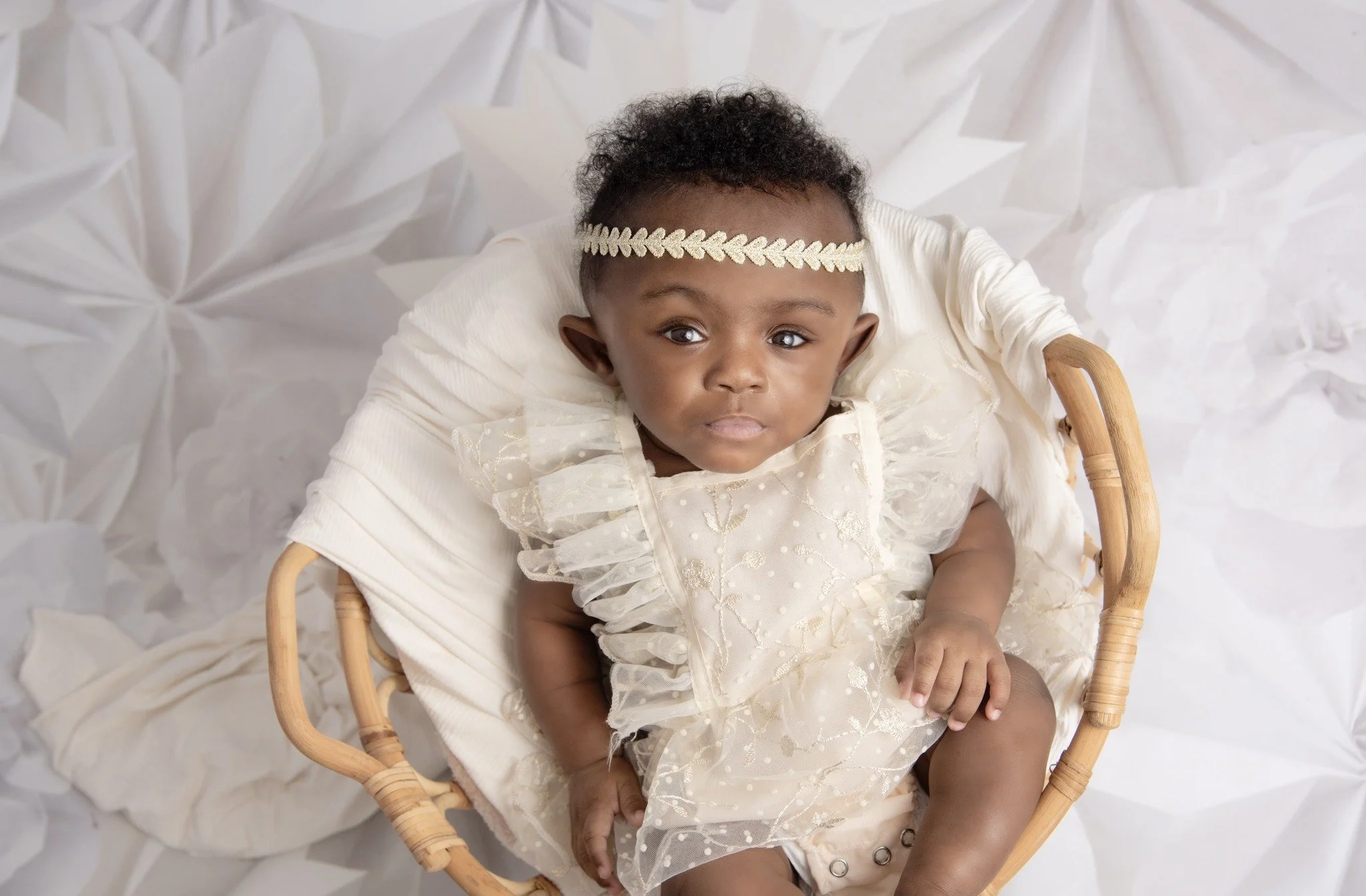 A baby girl wearing a cream-colored dress with ruffles and embroidery, sitting in a wicker basket, with a cream-colored headband with leaves, on a white textured background.