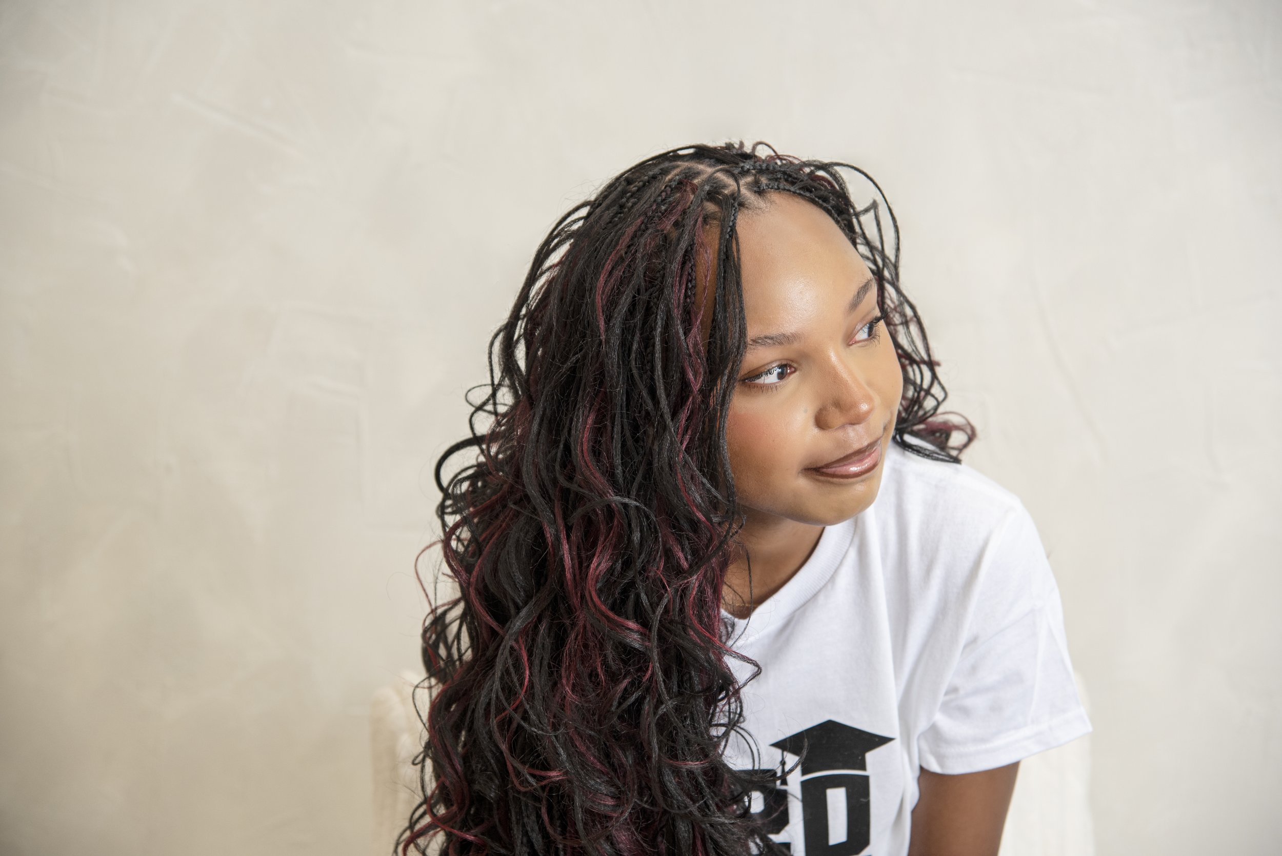 A young woman with long, wavy hair with reddish highlights, wearing a white t-shirt, looking to her right, against a plain light-colored wall.