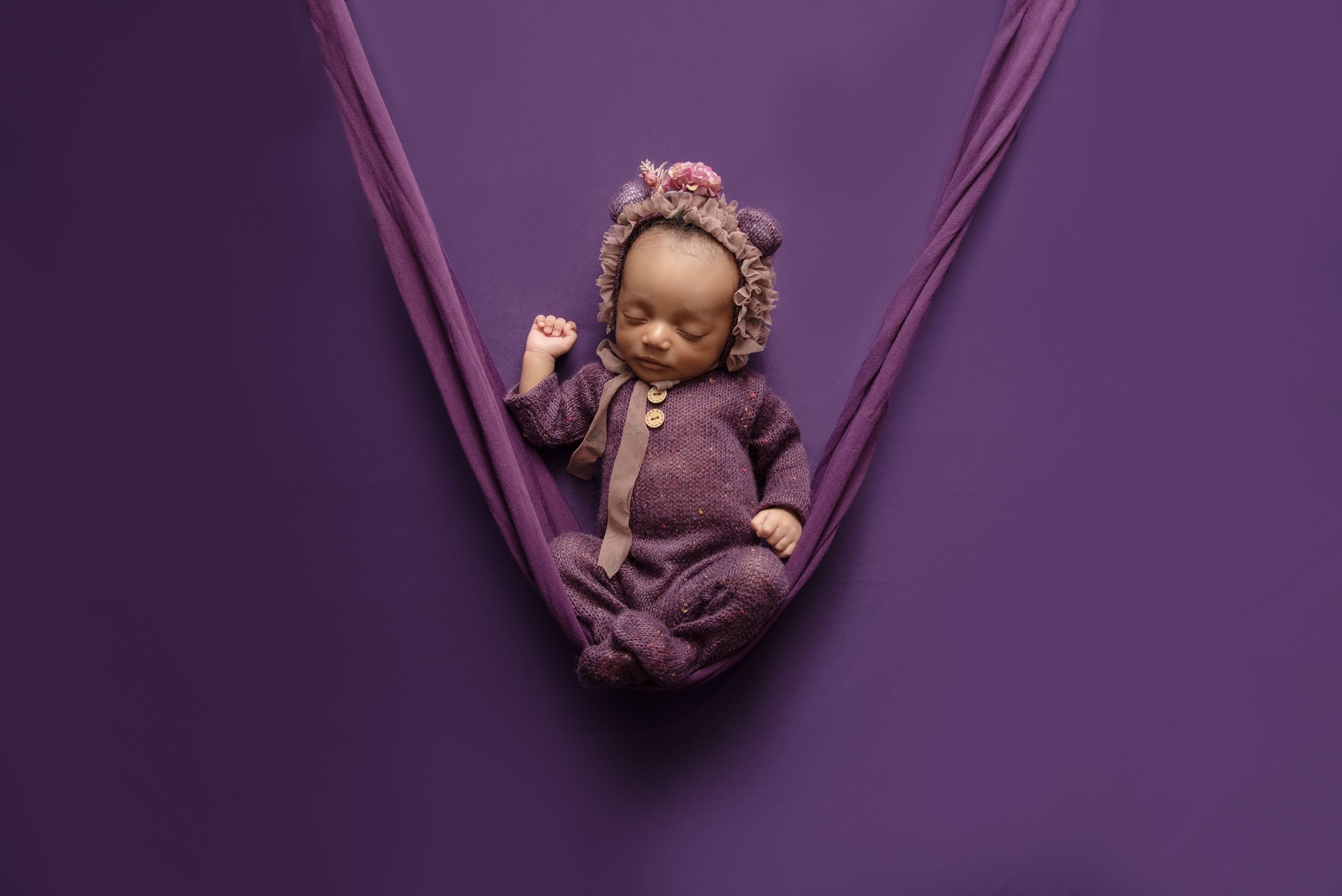 A sleeping baby dressed in purple pajamas, wearing a matching bonnet with flowers and bear ears, lying in a purple hammock against a purple background.