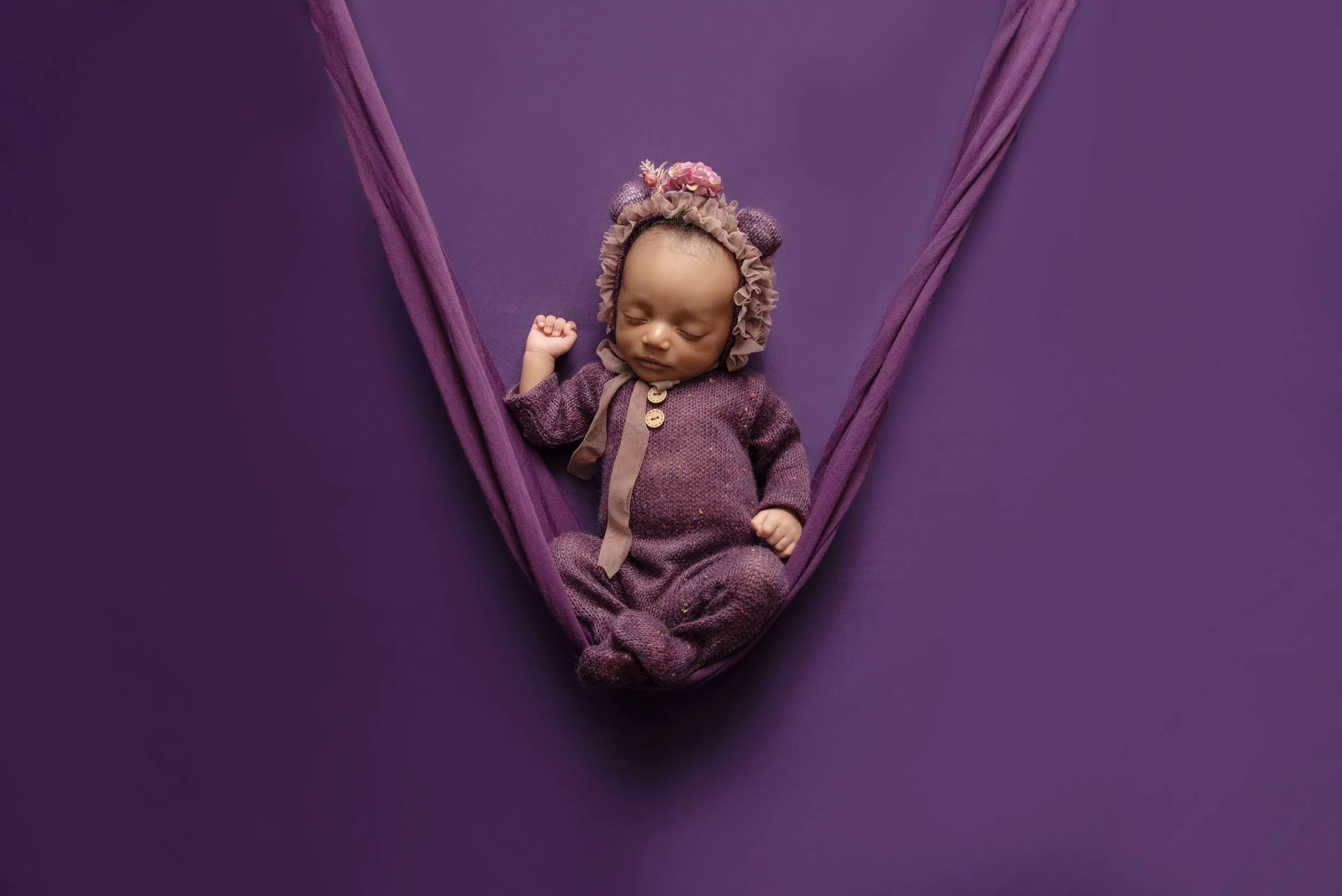 A sleeping baby dressed in purple pajamas, wearing a matching bonnet with flowers and bear ears, lying in a purple hammock against a purple background.