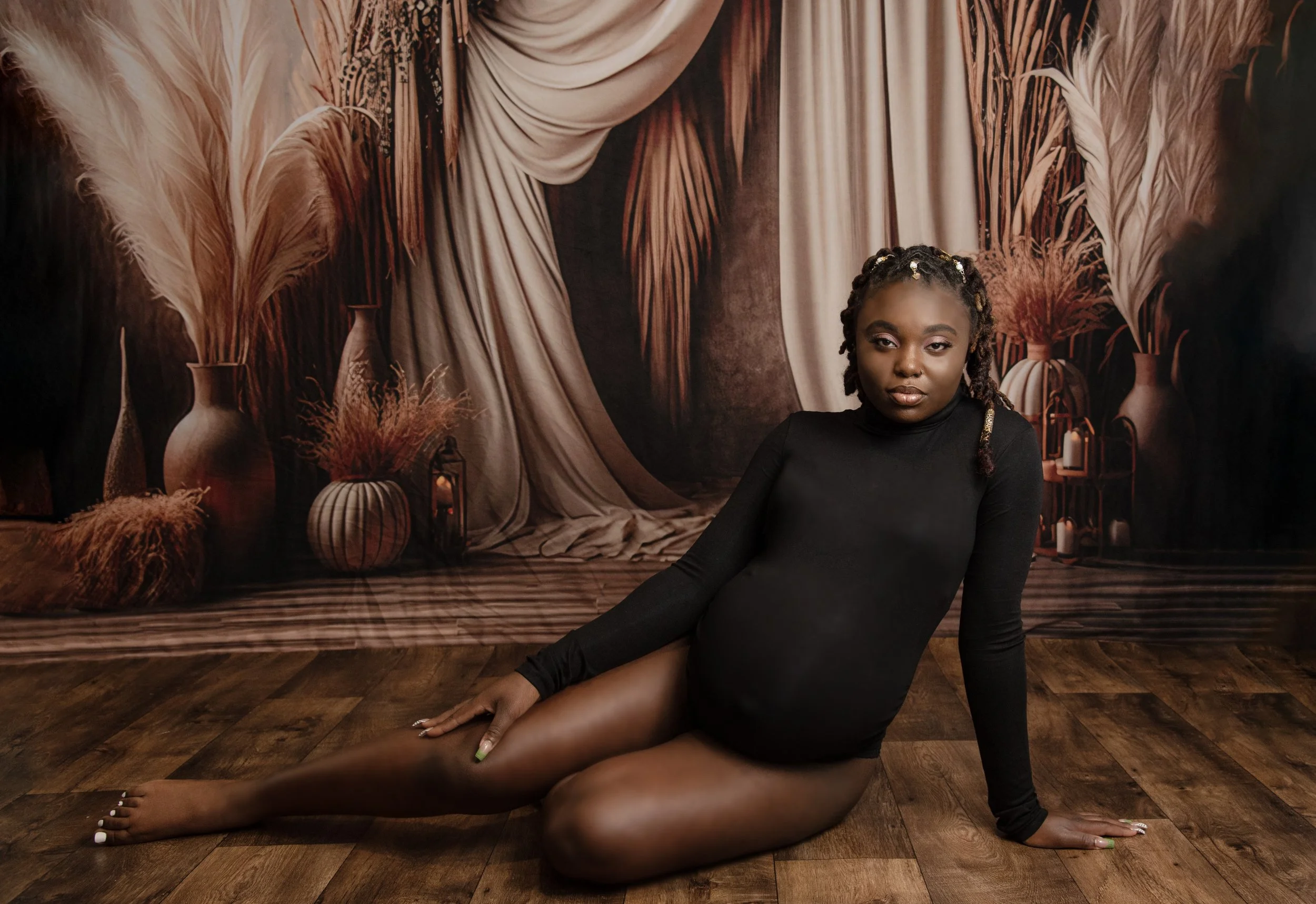 A young woman in a black long-sleeve top sitting on a wooden floor in front of a backdrop with silk curtains and tall vases with pampas grass, with some lanterns on the floor.