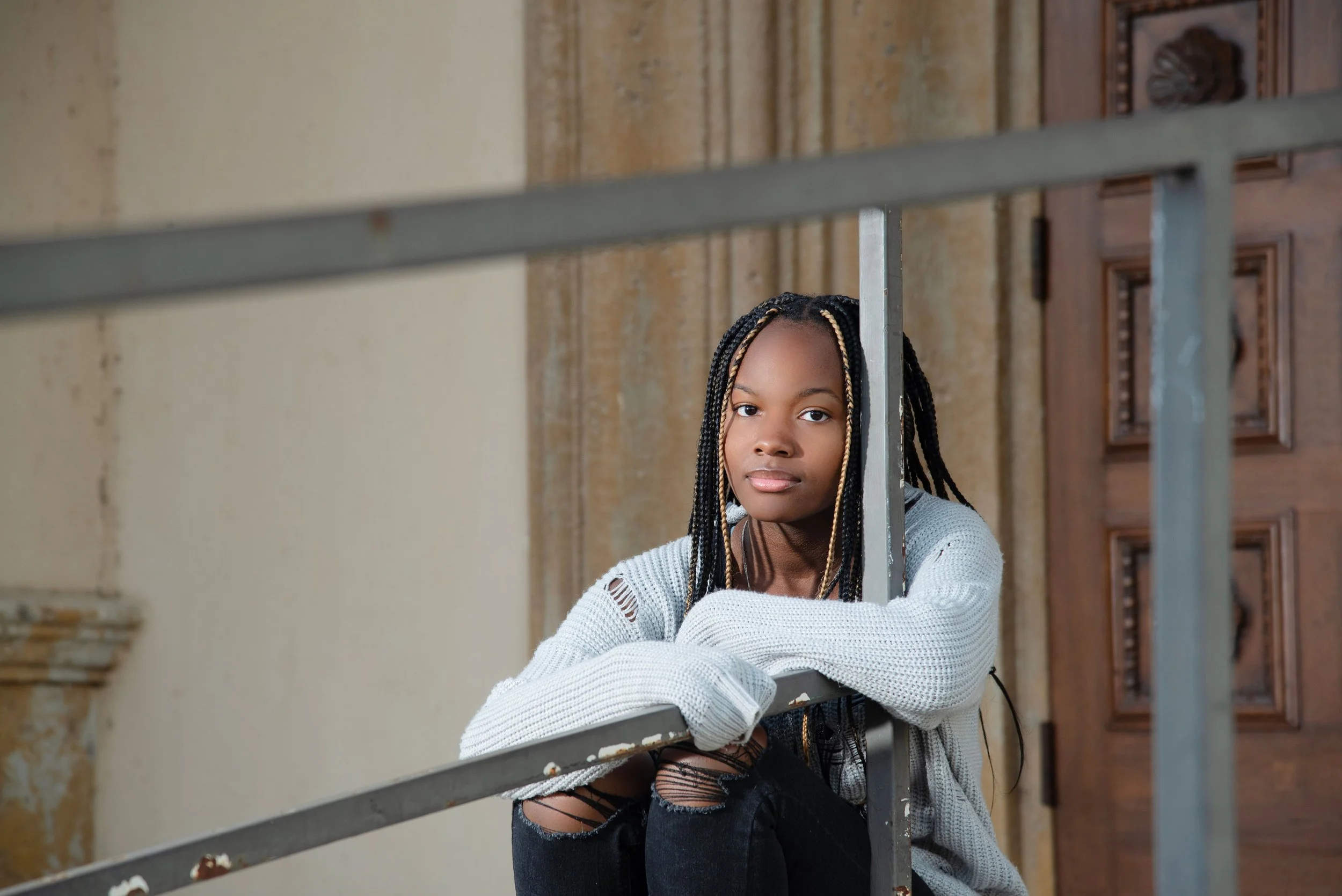 A young woman with braided hair sitting on a staircase in an old building, resting her arms on the metal railing.