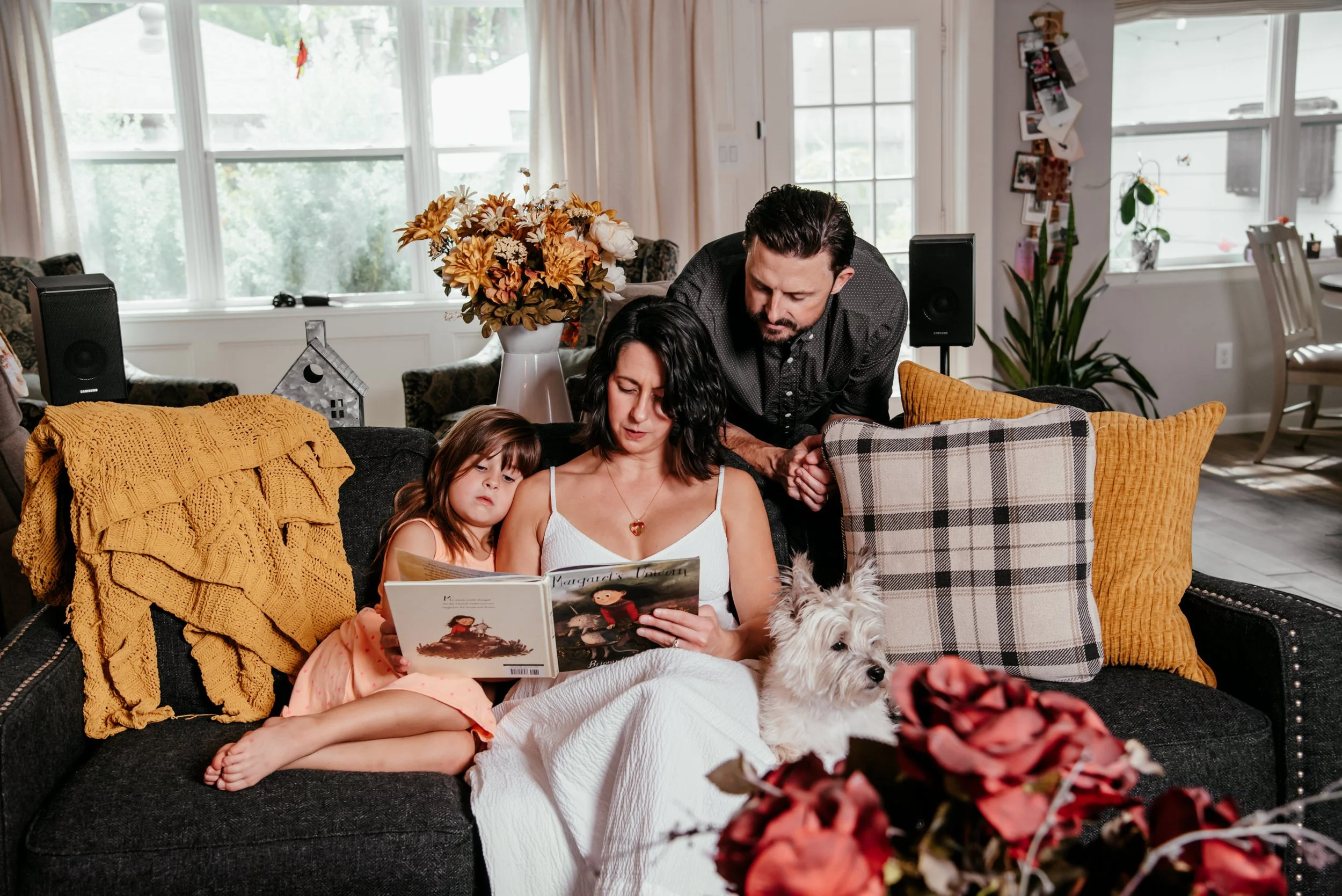A family sitting on a black couch in a living room, with a mother reading a book, a young girl lying beside her, and a man leaning over. There is a white dog sitting on the mother's lap. The room is decorated with flowers, pillows, and photos on the 