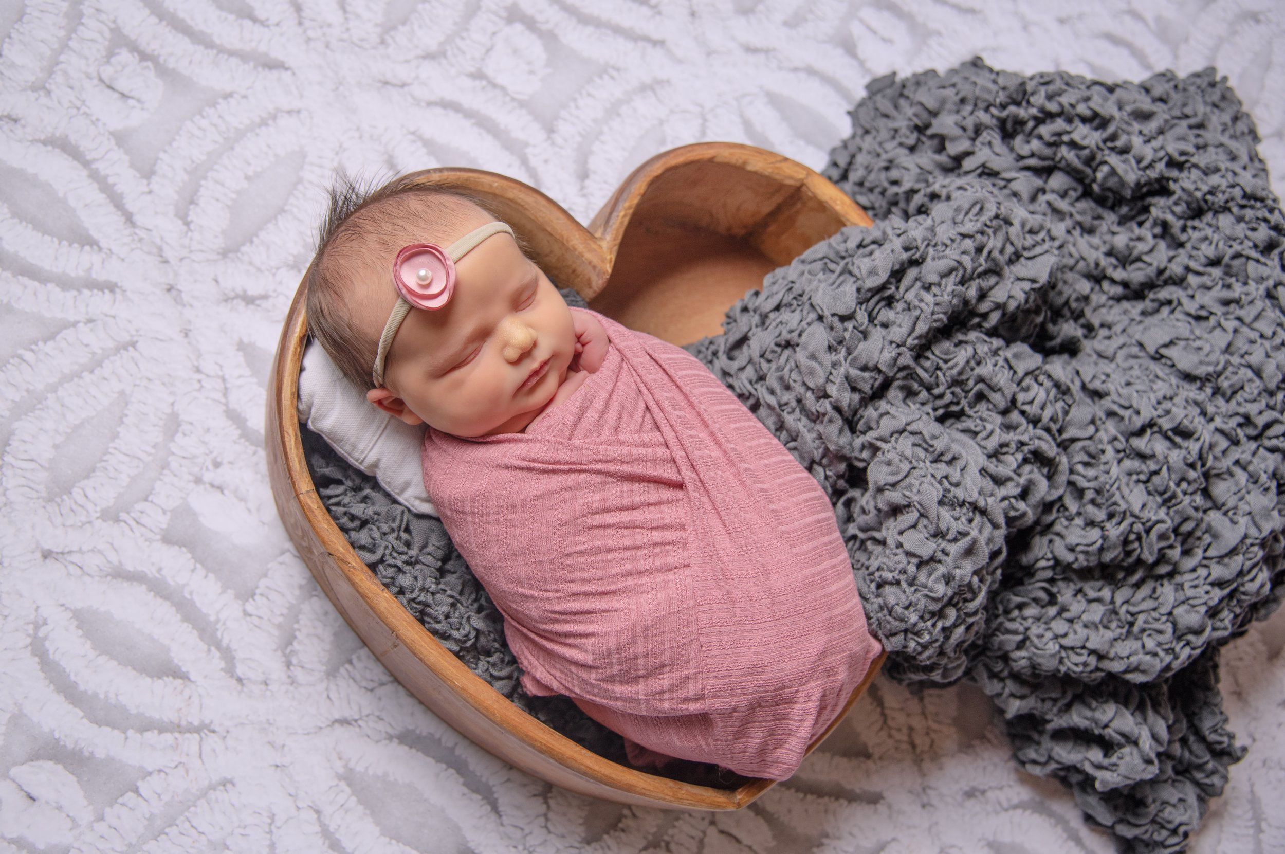 Newborn baby girl wrapped in pink on gray blanket in heart shaped prop against abstract backdrop