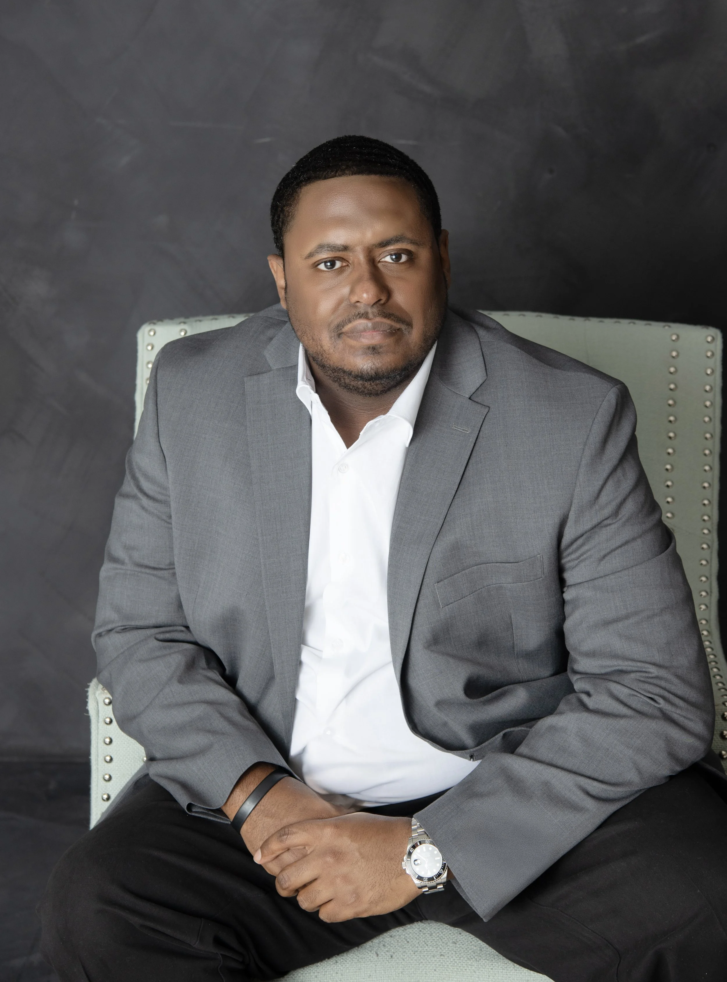 A man in a gray suit and white shirt sitting on a light-colored chair against a dark background for corporate headshot.