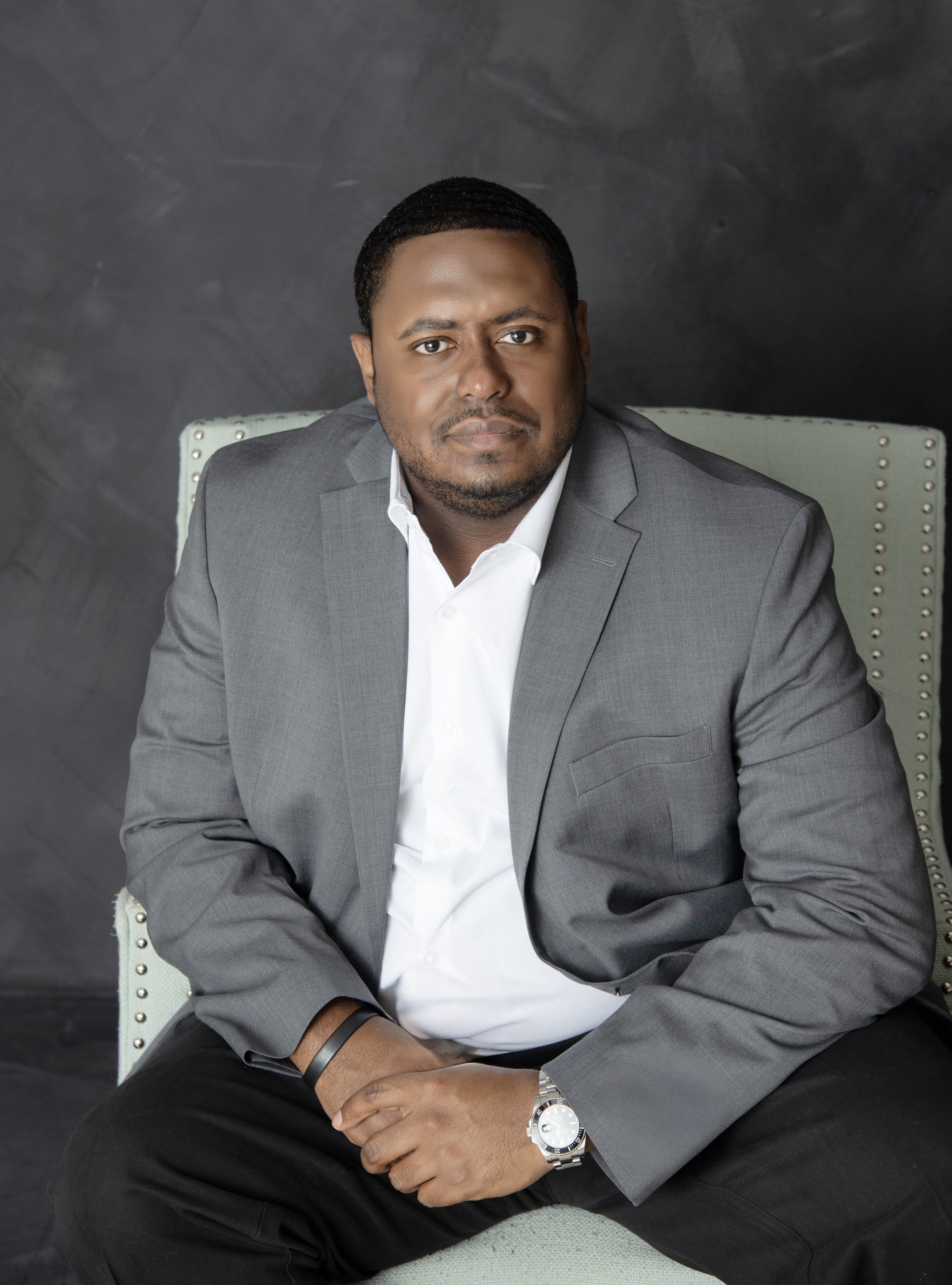 A man in a gray suit and white shirt sitting on a light-colored chair against a dark background for corporate headshot.