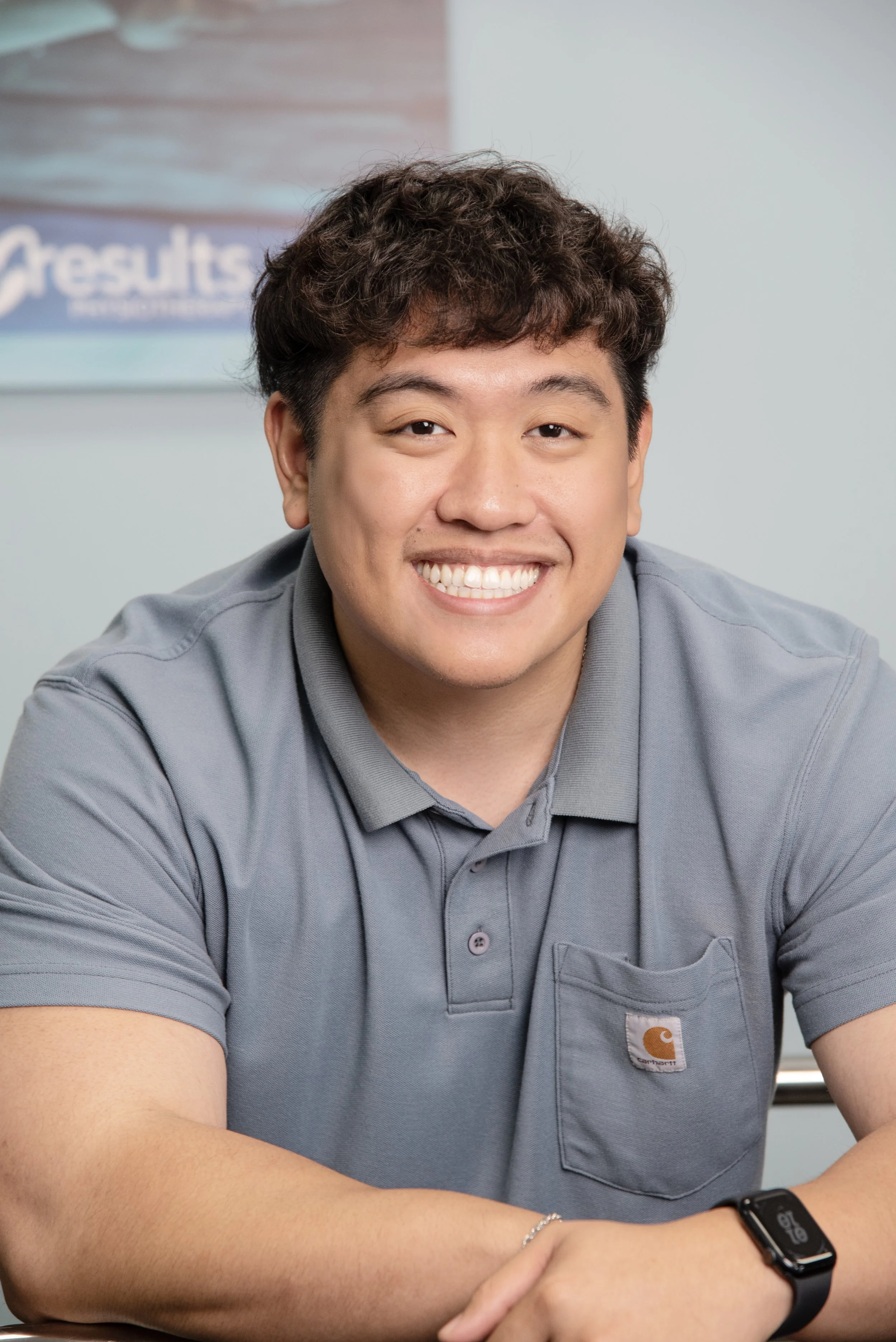 Young male therapy assistant smiling, wearing a gray polo shirt in front of a poster at Results Physiotherapy.