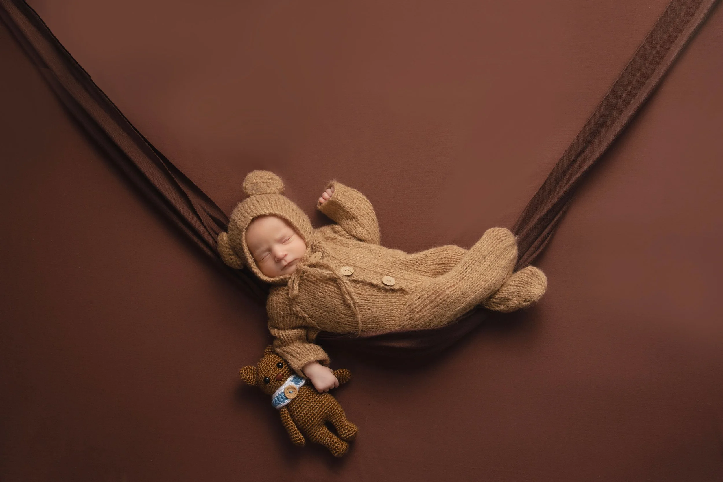 Newborn baby girl in teddy bear outfit holding teddy bear on on brown background