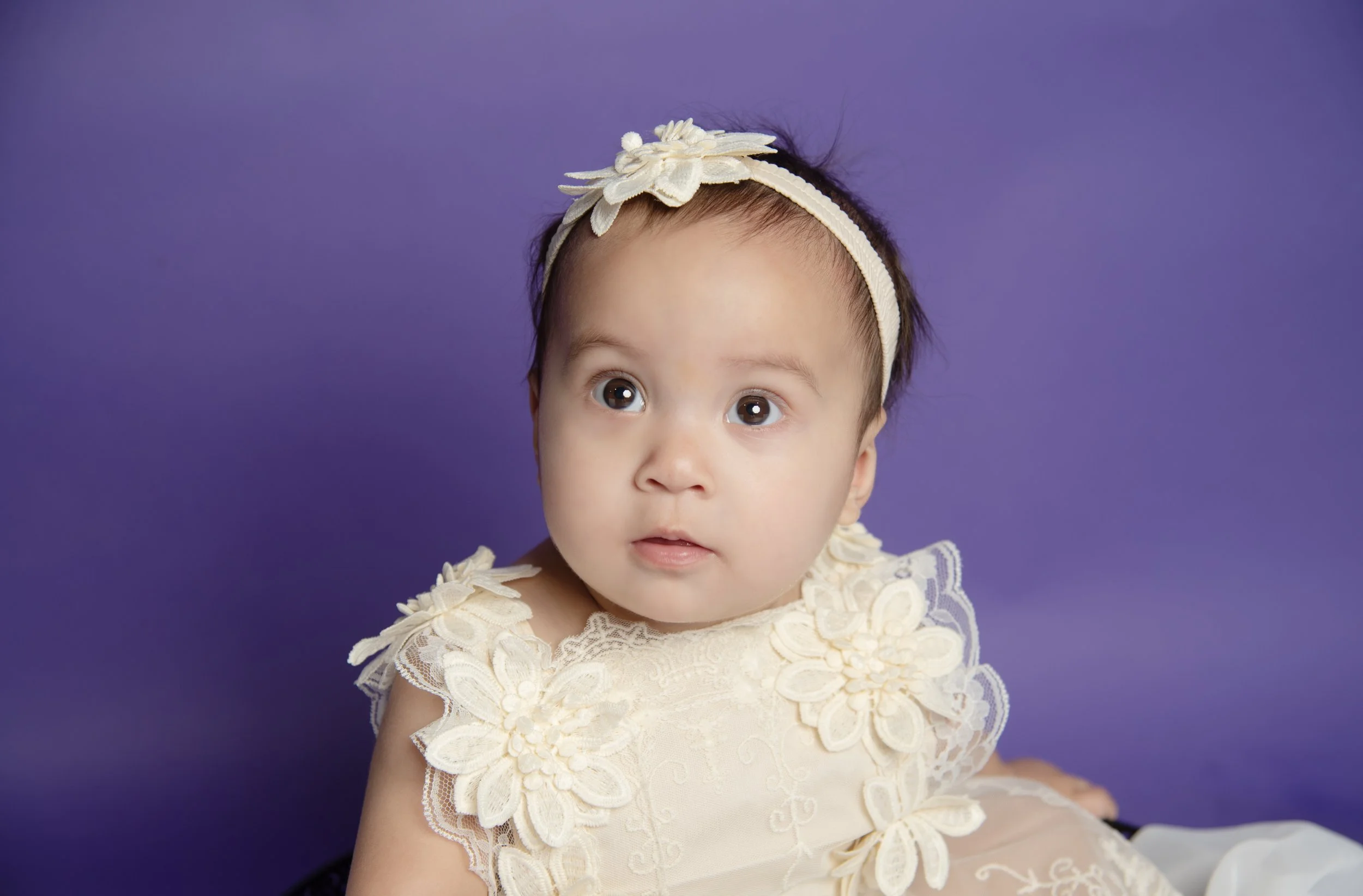 Young girl with big brown eyes and dark hair wearing a cream-colored dress with floral lace appliqué and a matching headband against a purple background.