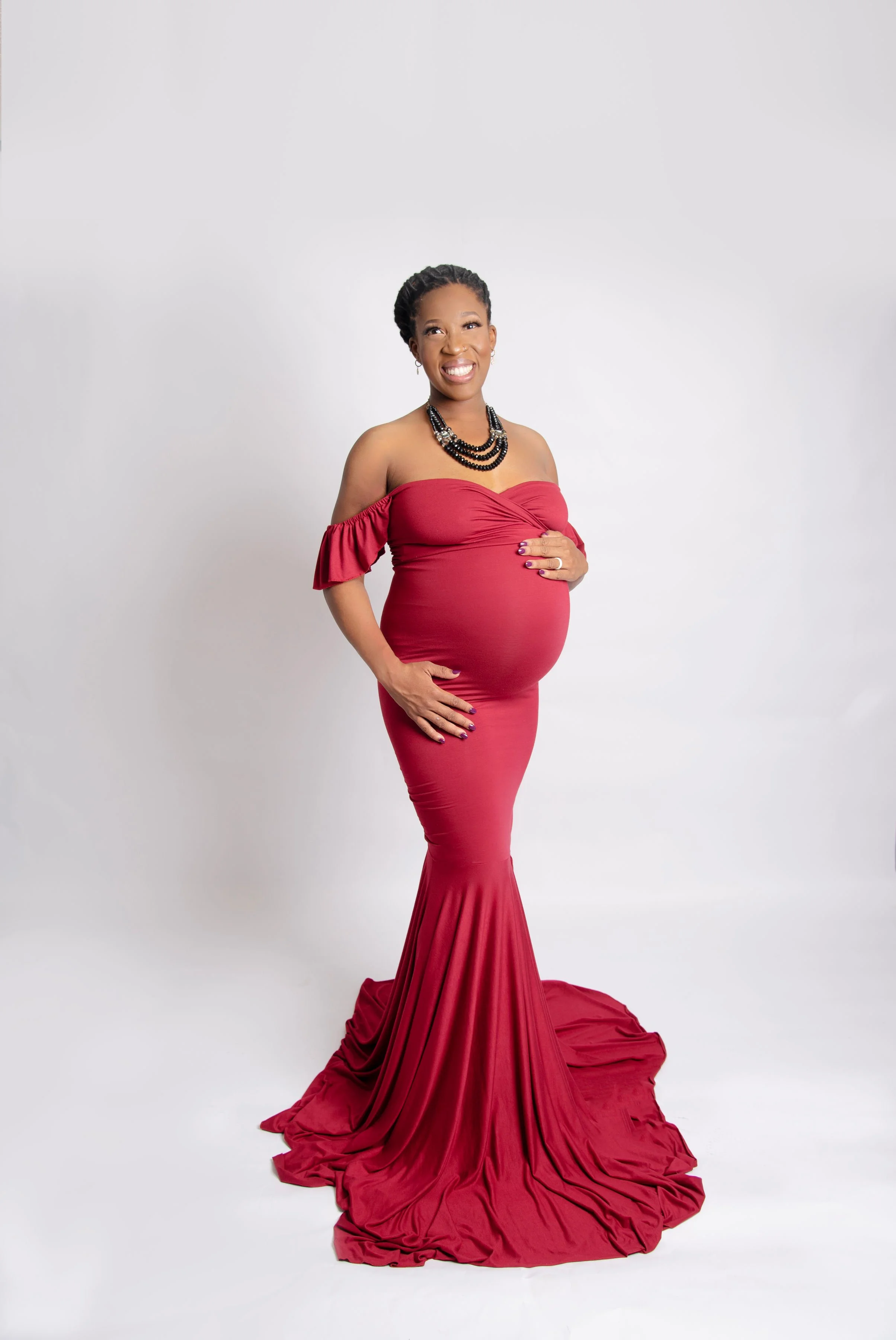 A pregnant woman in a red off-shoulder gown with ruched bust and a long, flowing train, standing against a plain white background, smiling and touching her belly.