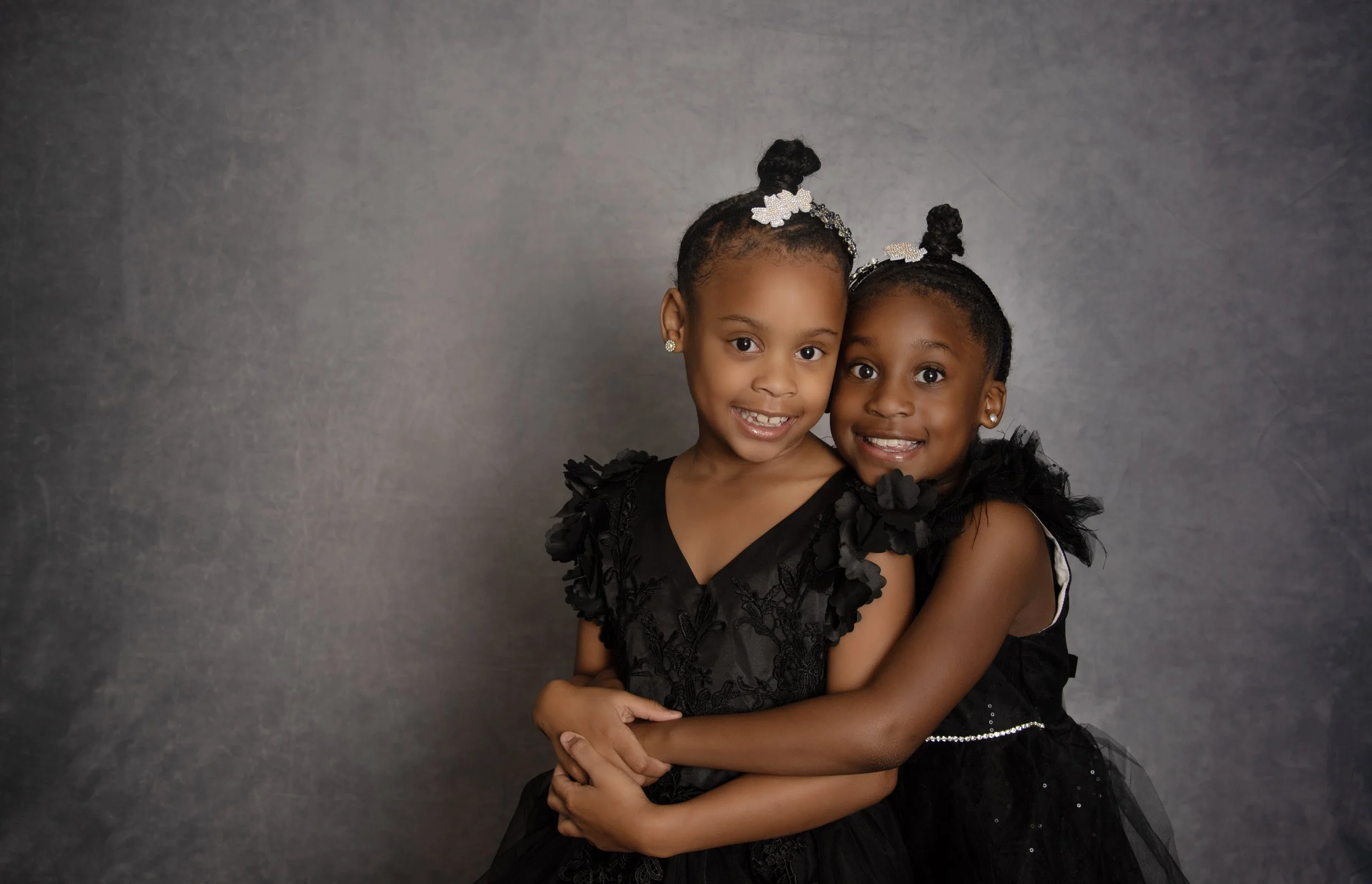 Two young girls wearing black dresses with floral embellishments, smiling, standing close together against a gray backdrop, hugging.