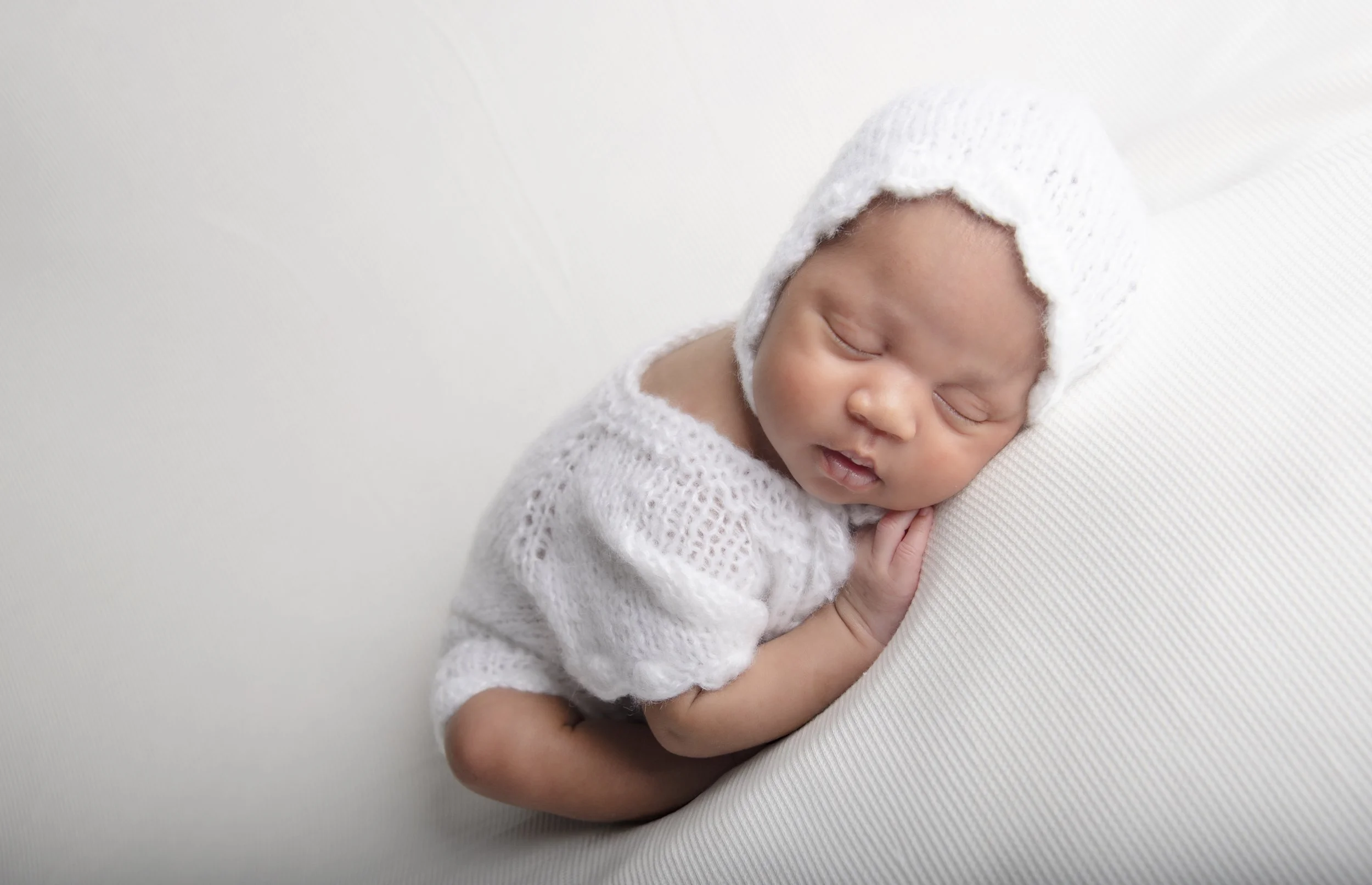 Sleeping newborn baby wearing a white knit hat and sweater, lying on their side on a white textured surface.