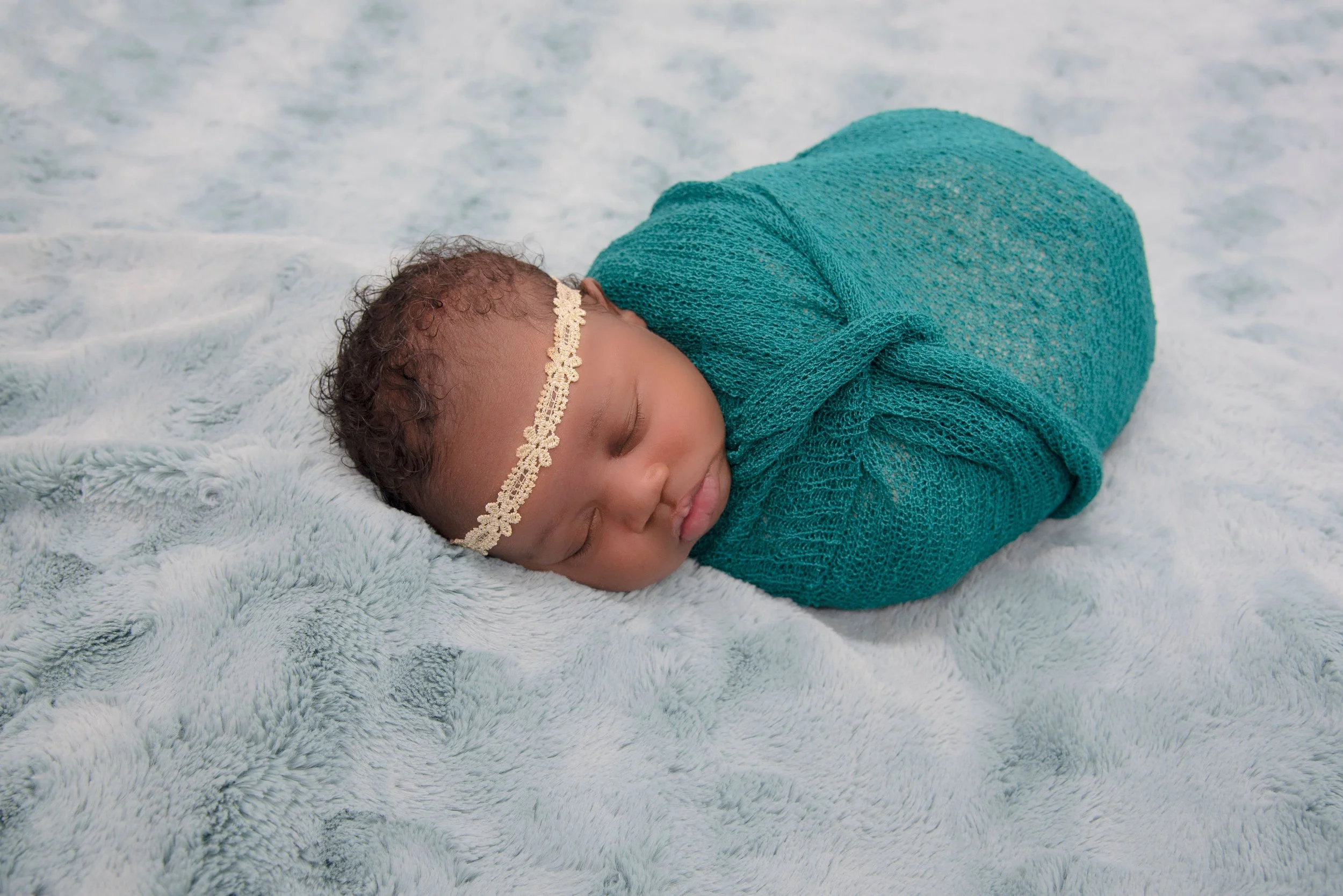 A newborn baby wrapped in a teal knitted blanket, sleeping on a soft, light-colored plush surface, wearing a lace headband.