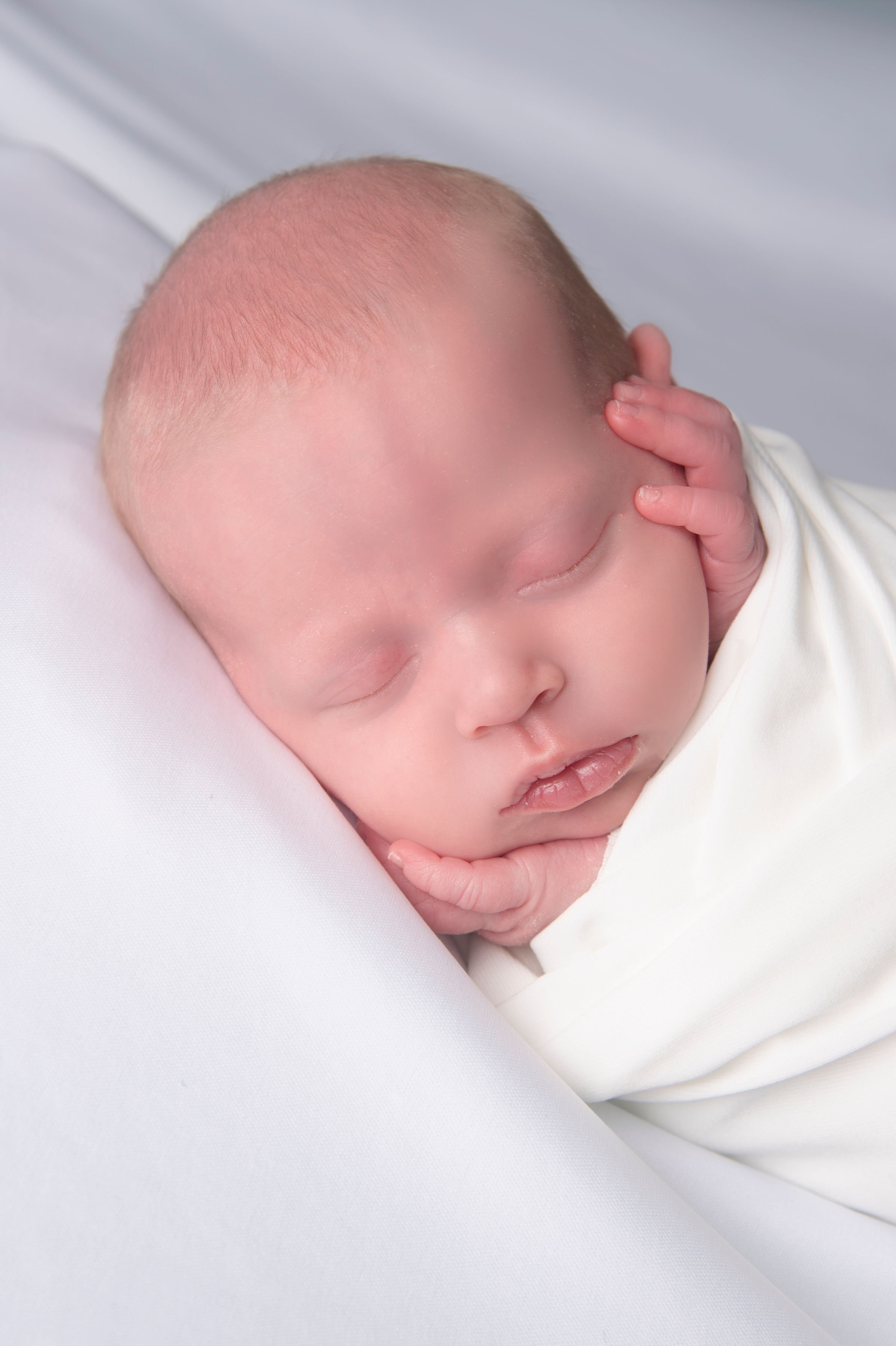 A sleeping baby resting on a white pillow, with eyes closed and one hand gently touching its face.