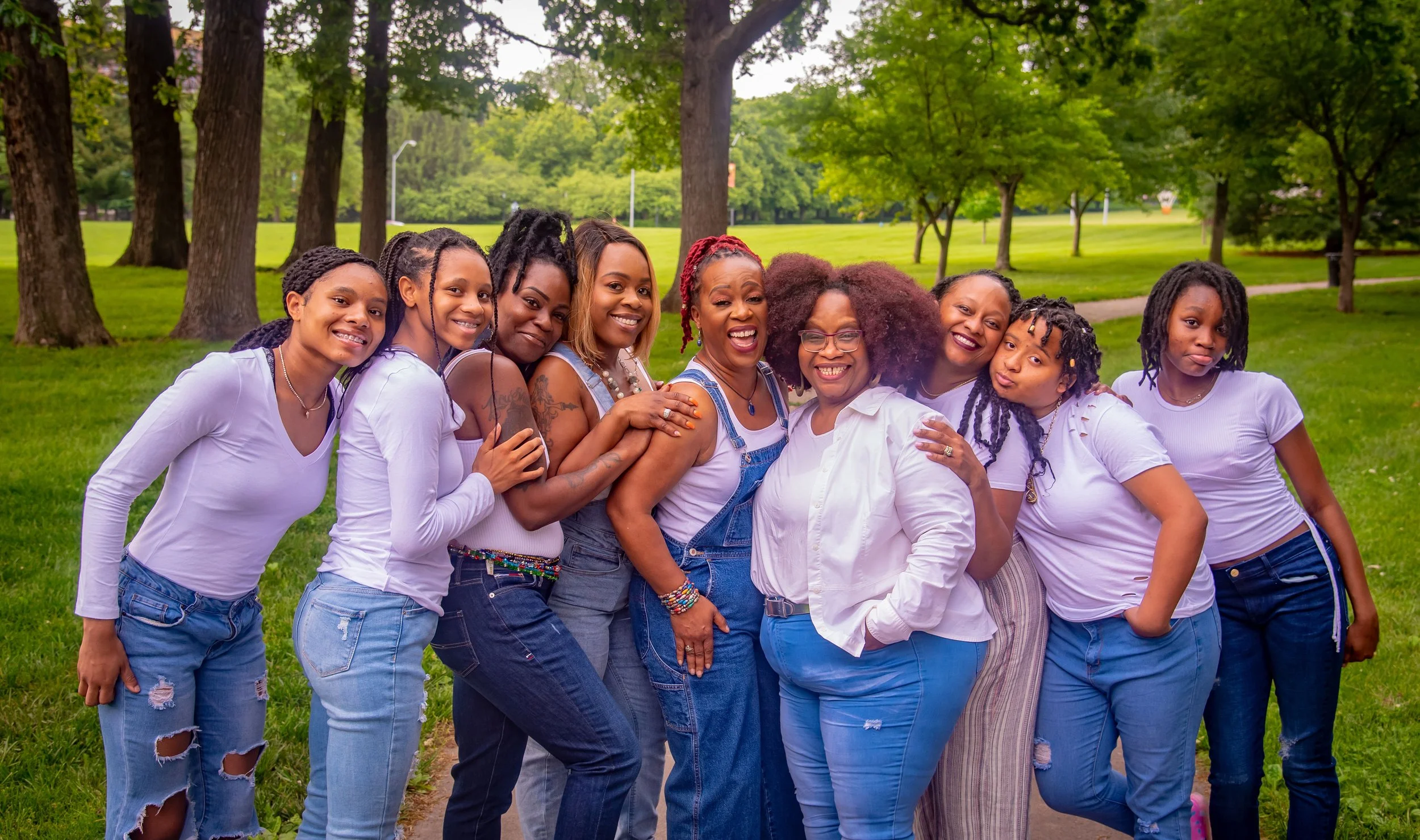 Group of ten women of varying ages smiling and embracing outdoors in a park with trees and grass.