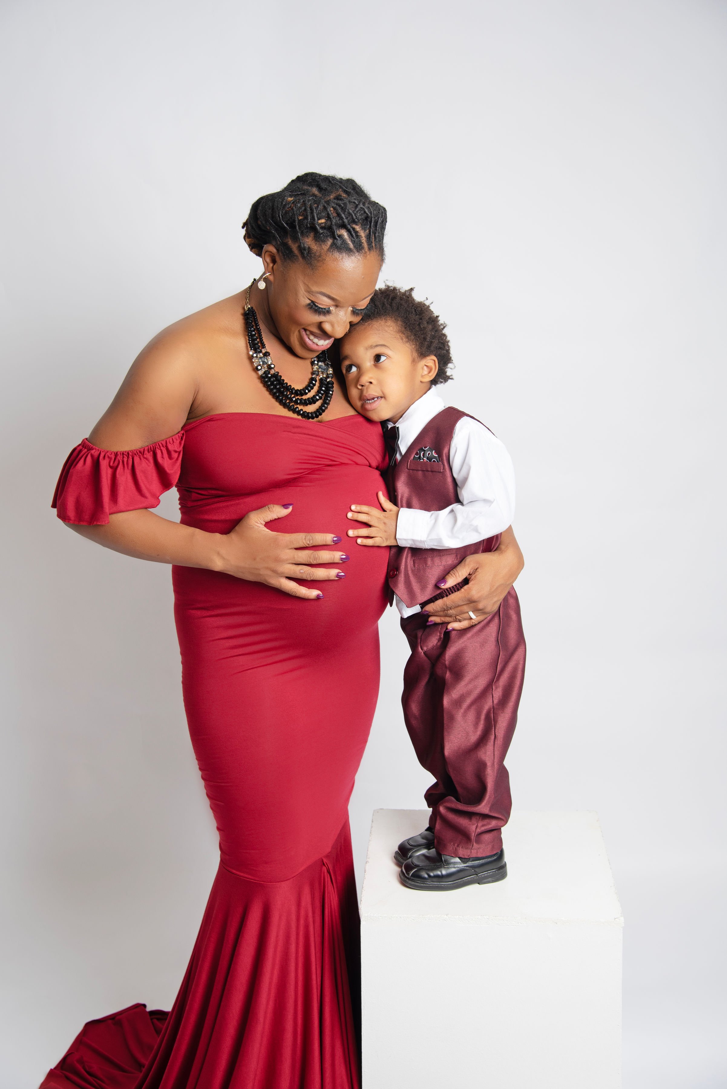 A woman in a red off-shoulder dress and a young boy in formal attire are smiling and hugging, with the boy standing on a white platform.