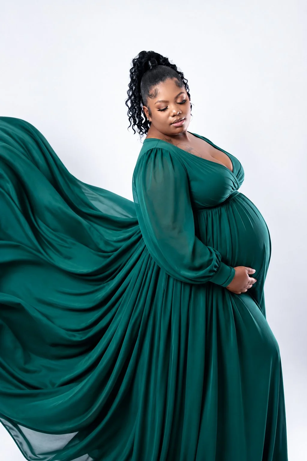 A pregnant woman in a flowing emerald green dress with long sleeves, standing against a plain white background, with her eyes closed and her hand resting on her baby bump.