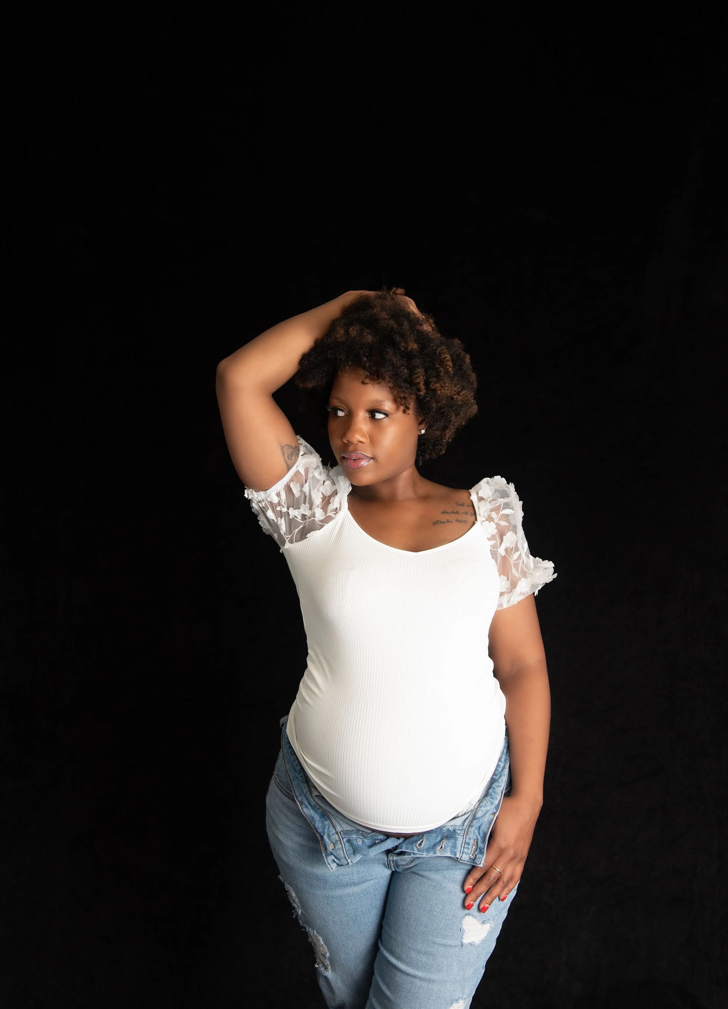A pregnant woman with curly hair stands against a black background, wearing a white top with lace sleeves and distressed jeans, with her right hand resting on her head.