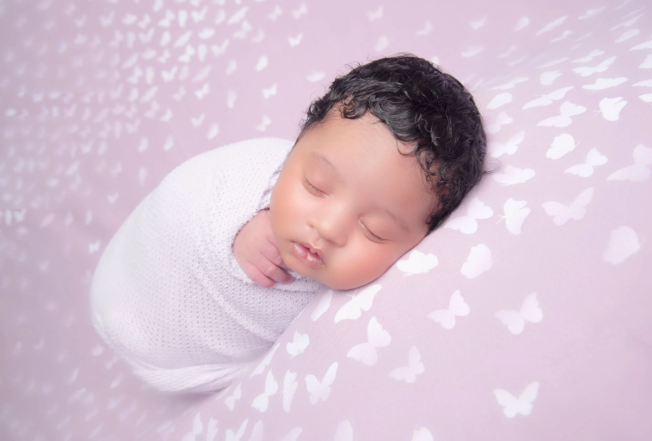 A sleeping baby with curly black hair wrapped in a white blanket, lying on a pink surface with white butterfly patterns.