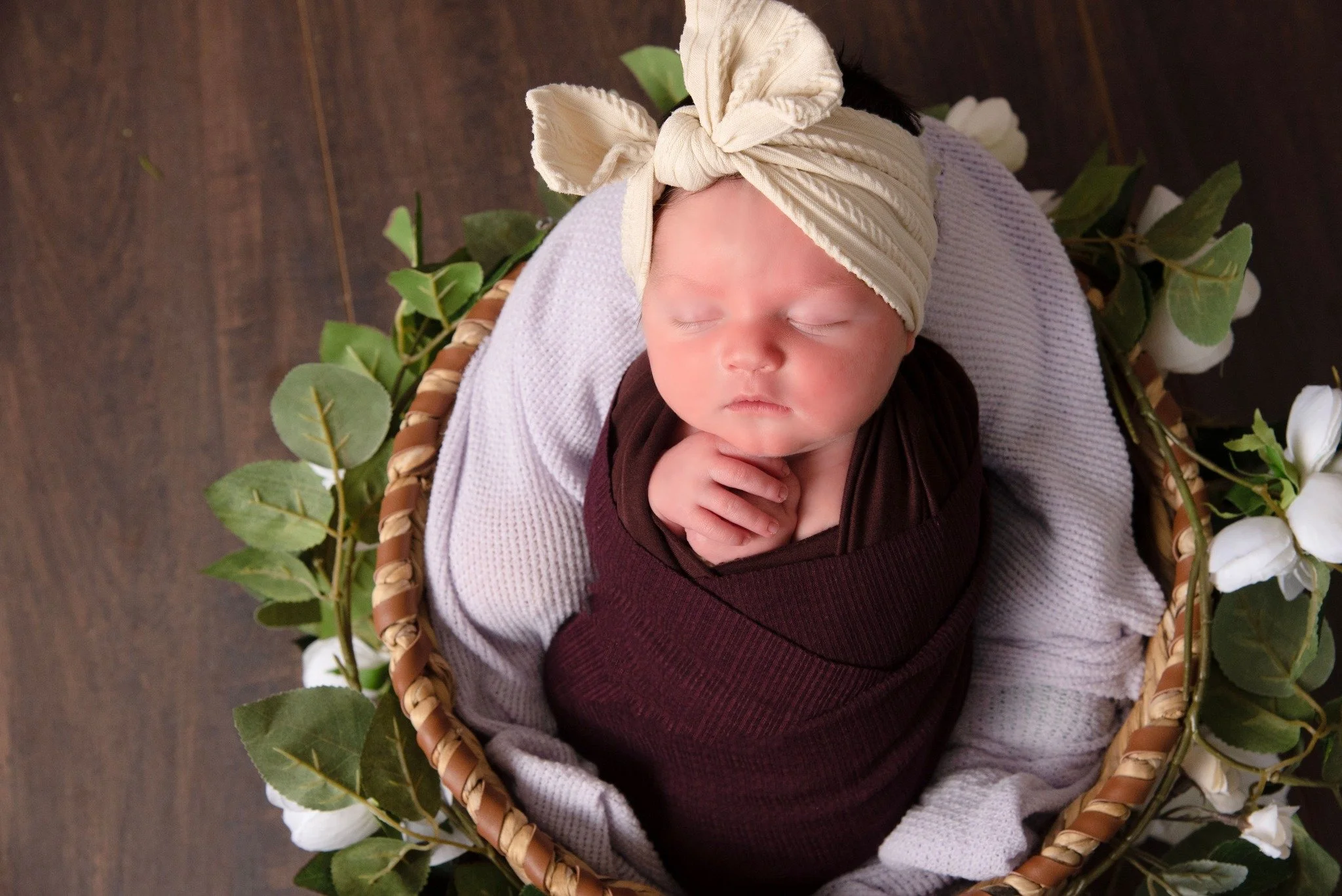 A sleeping baby in a brown swaddle, wearing a cream-colored headband with a bow, lying in a basket decorated with green leaves and white flowers on a wooden floor.
