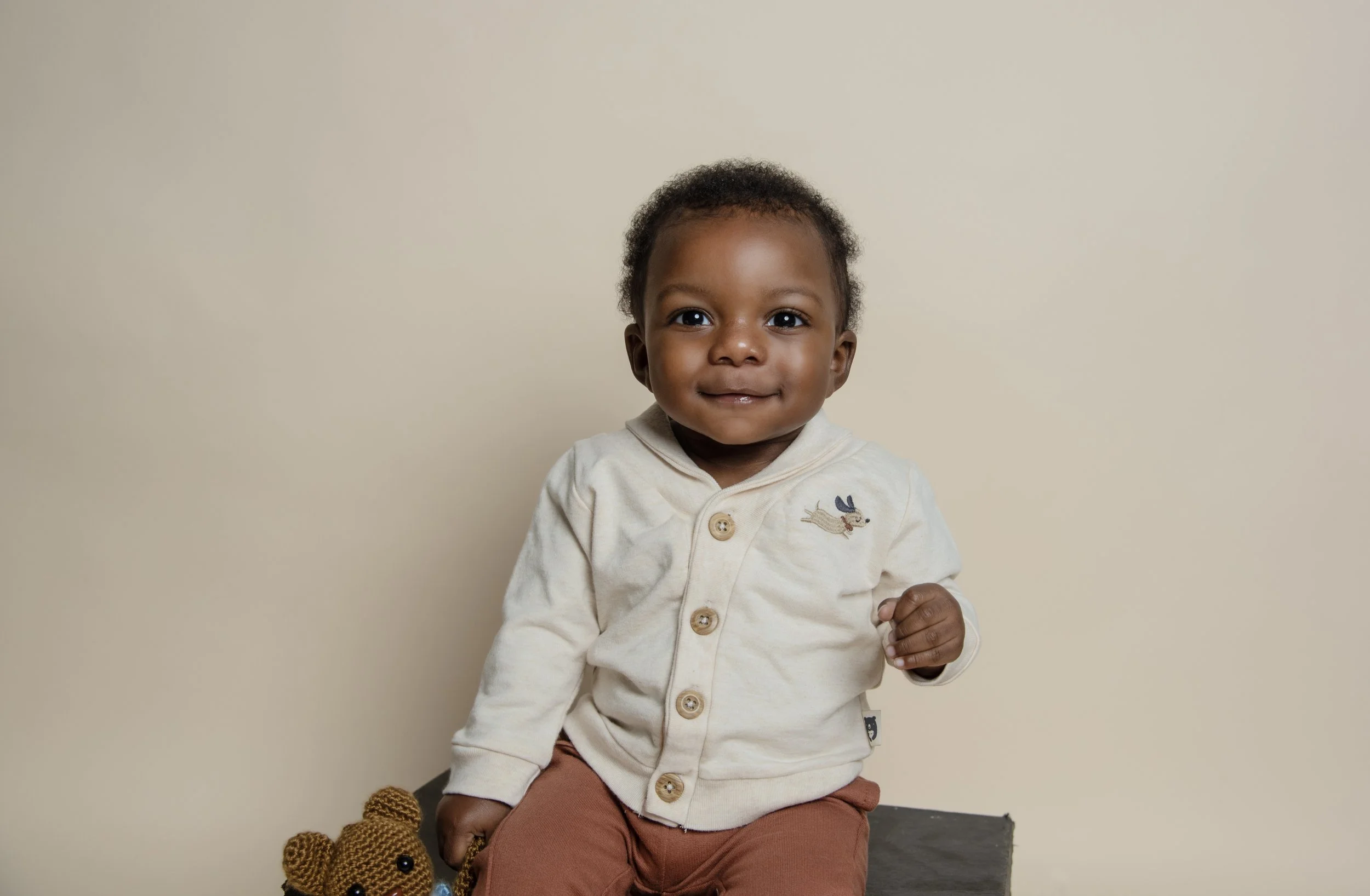 A young child with dark skin and short curly hair sitting against a beige wall, wearing a cream-colored button-up shirt with a small dog embroidery on the chest and brown pants, holding a knitted teddy bear.