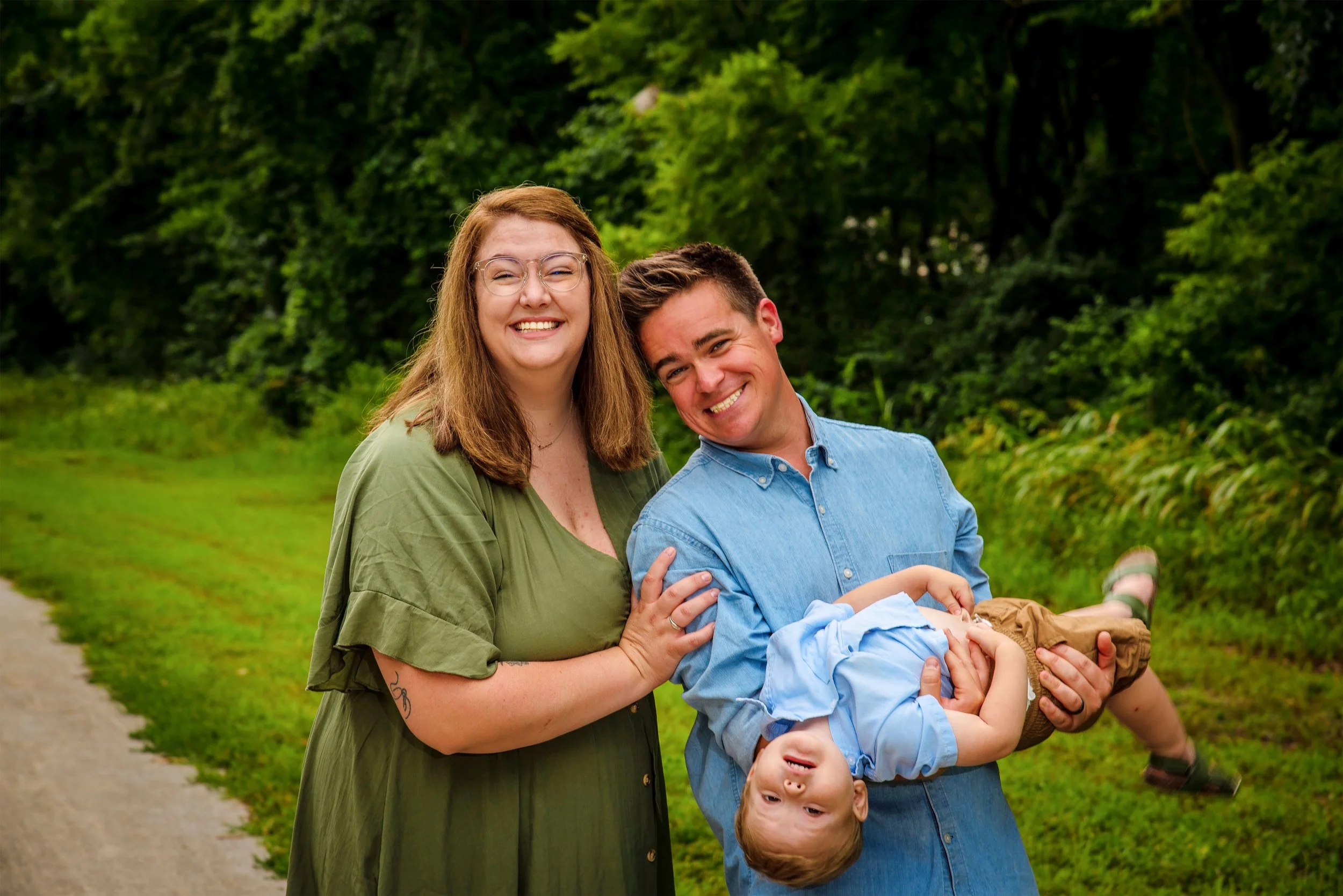 A happy family of three standing outdoors on a grassy area with trees in the background: a woman with red hair and glasses, a man with short dark hair, and a young boy being held upside down, all smiling.